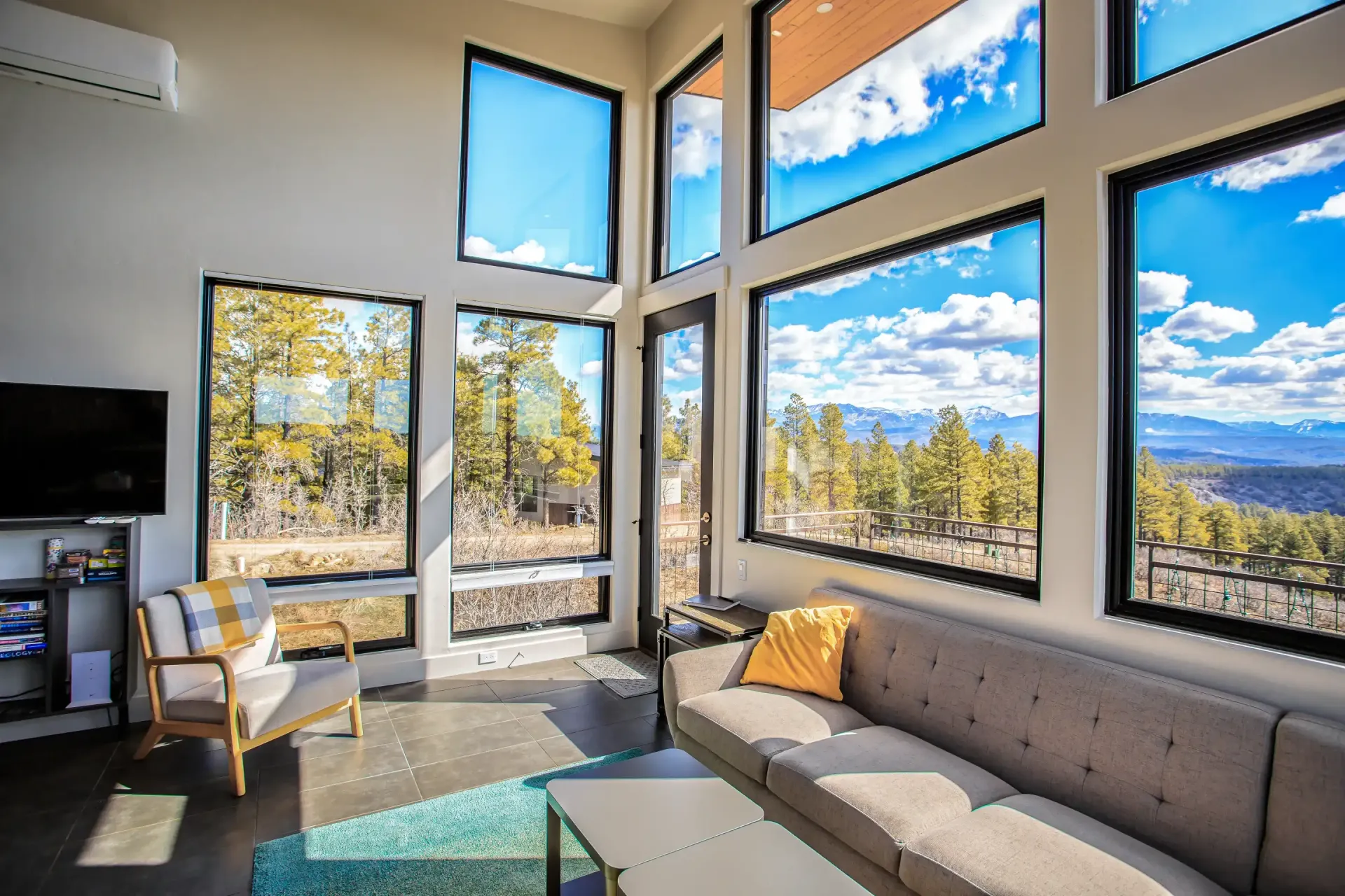 A bright living room with a grey sofa, armchair, and floor-to-ceiling windows showing a view of trees and mountains.