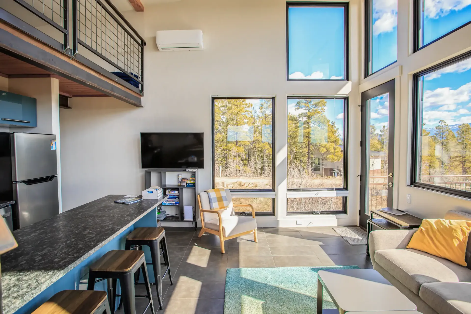A bright modern living room with a loft, kitchen island, stools, sofa, and large windows overlooking a wooded area.