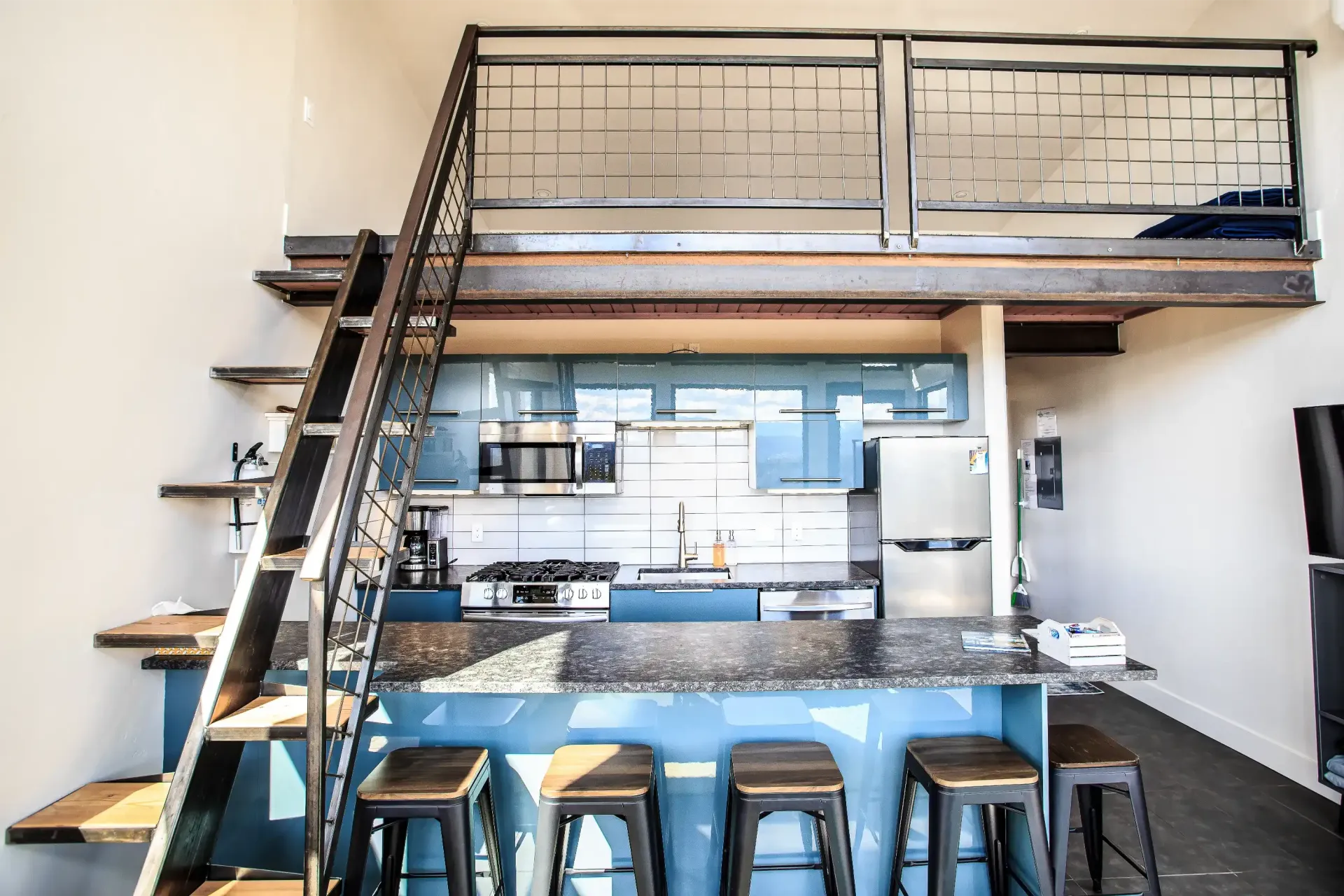 A kitchen with blue cabinetry and a granite island with four barstools, situated beneath a metal loft with a staircase.