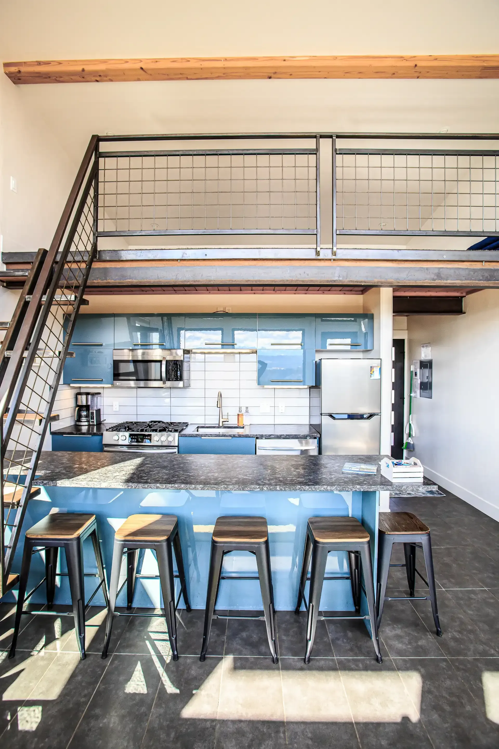 Modern kitchen with blue cabinetry, dark stone counters, four metal stools, and a metal staircase leading to a loft area.