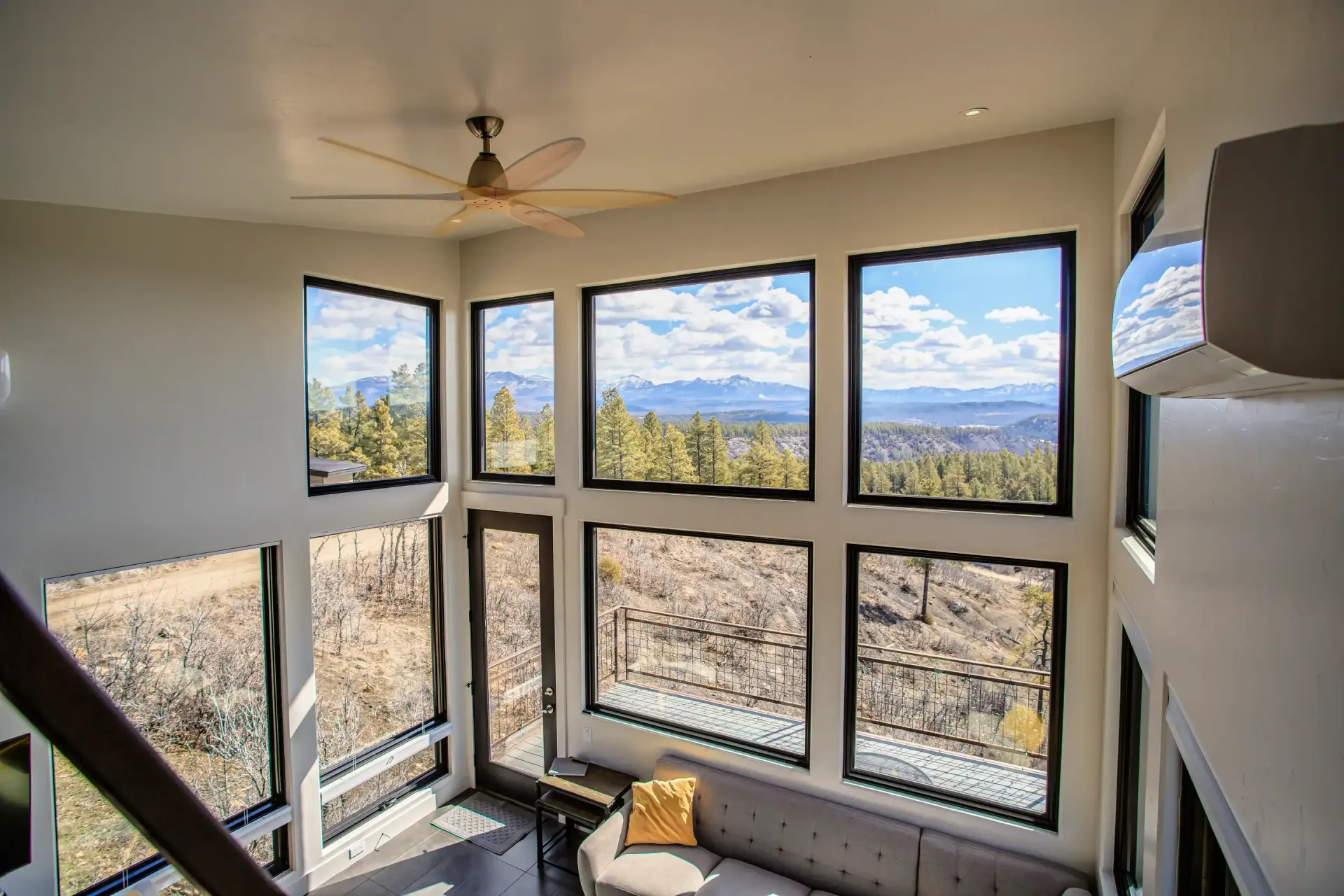 A modern living room with large windows showcasing mountain views, a grey sofa, and a ceiling fan.