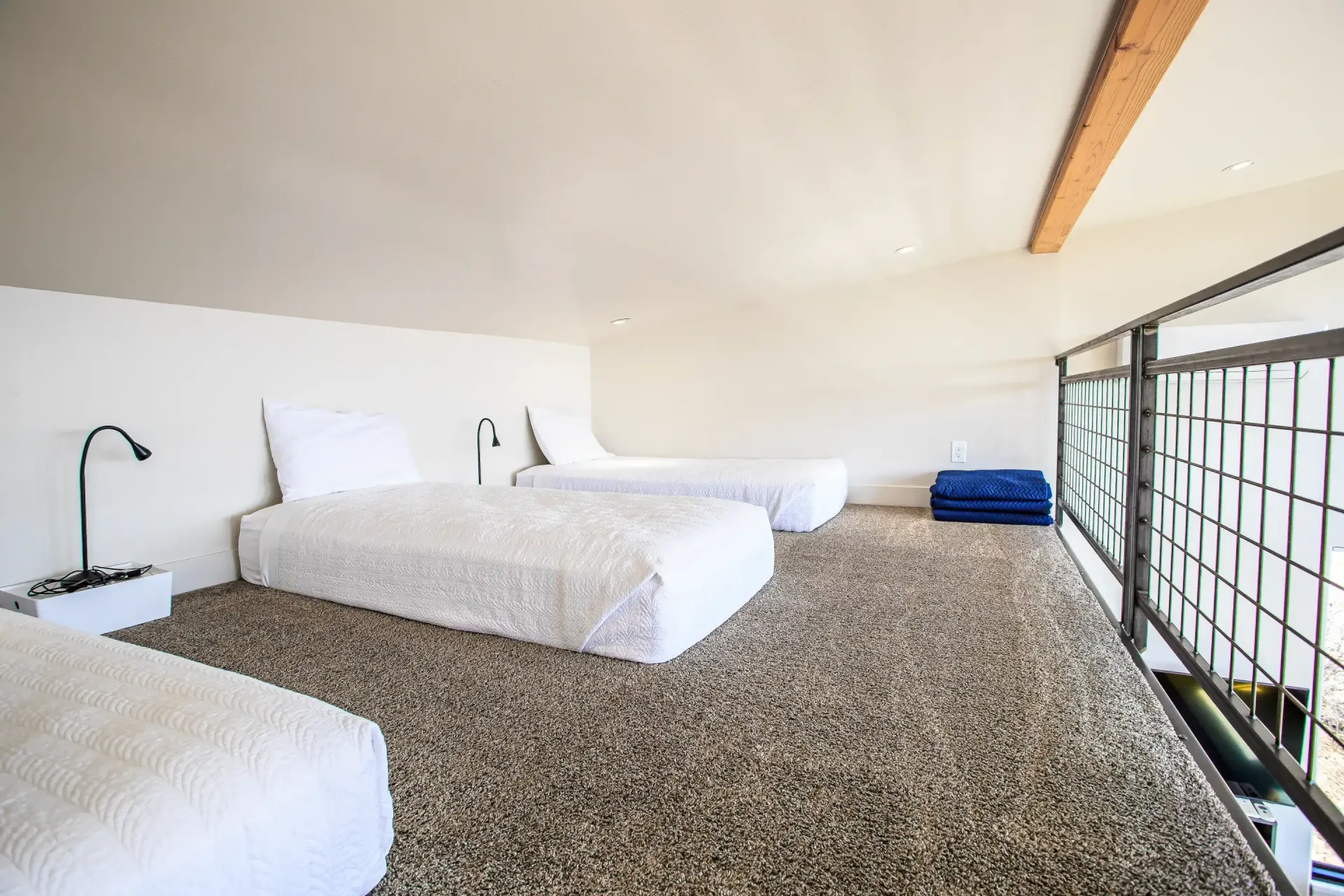 A loft space with three white beds on a textured carpeted floor, adjacent to a metal railing overlooking a lower level.