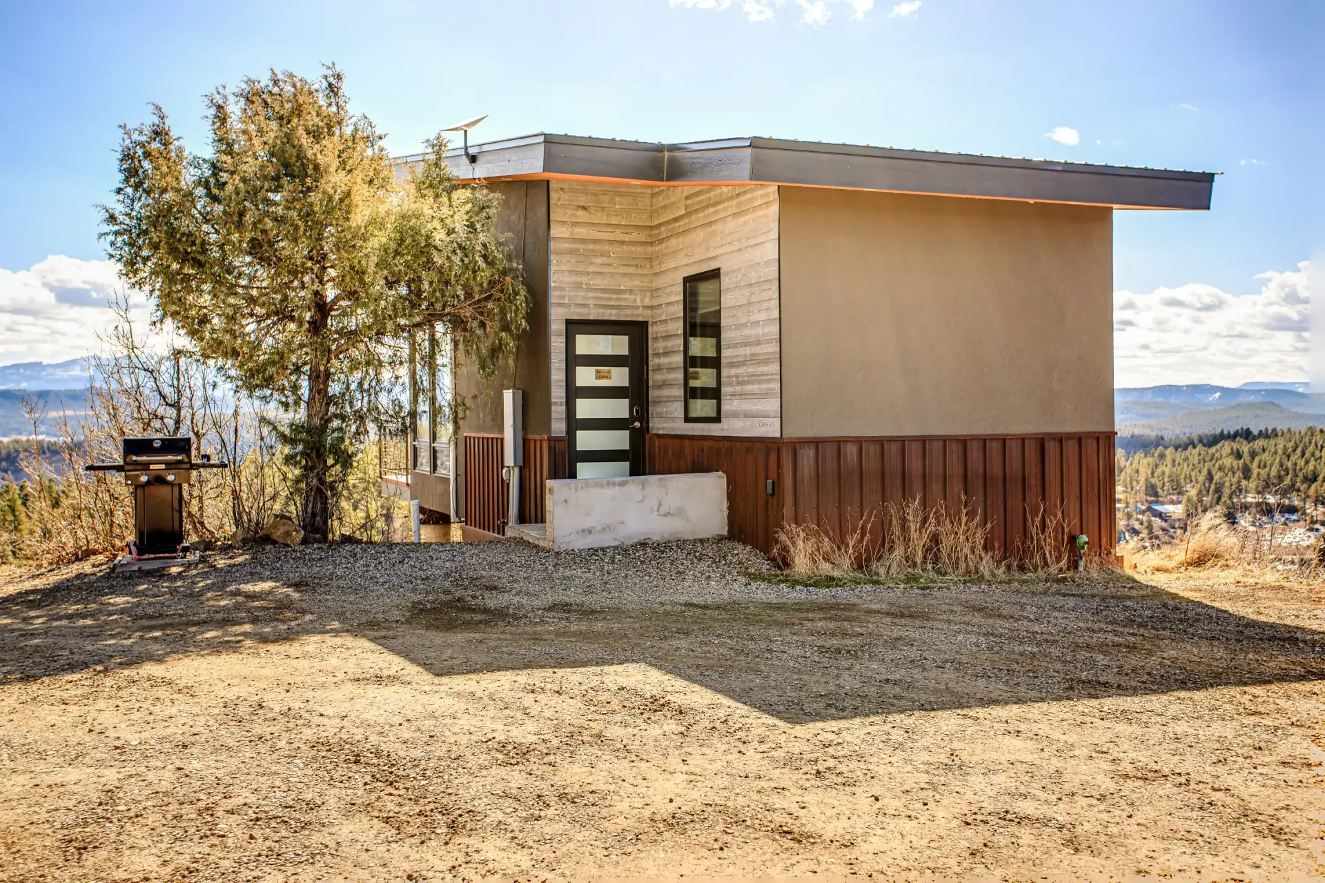 Modern cabin with stone and wood siding, a flat roof, and a front door, situated in a dry, mountainous landscape.