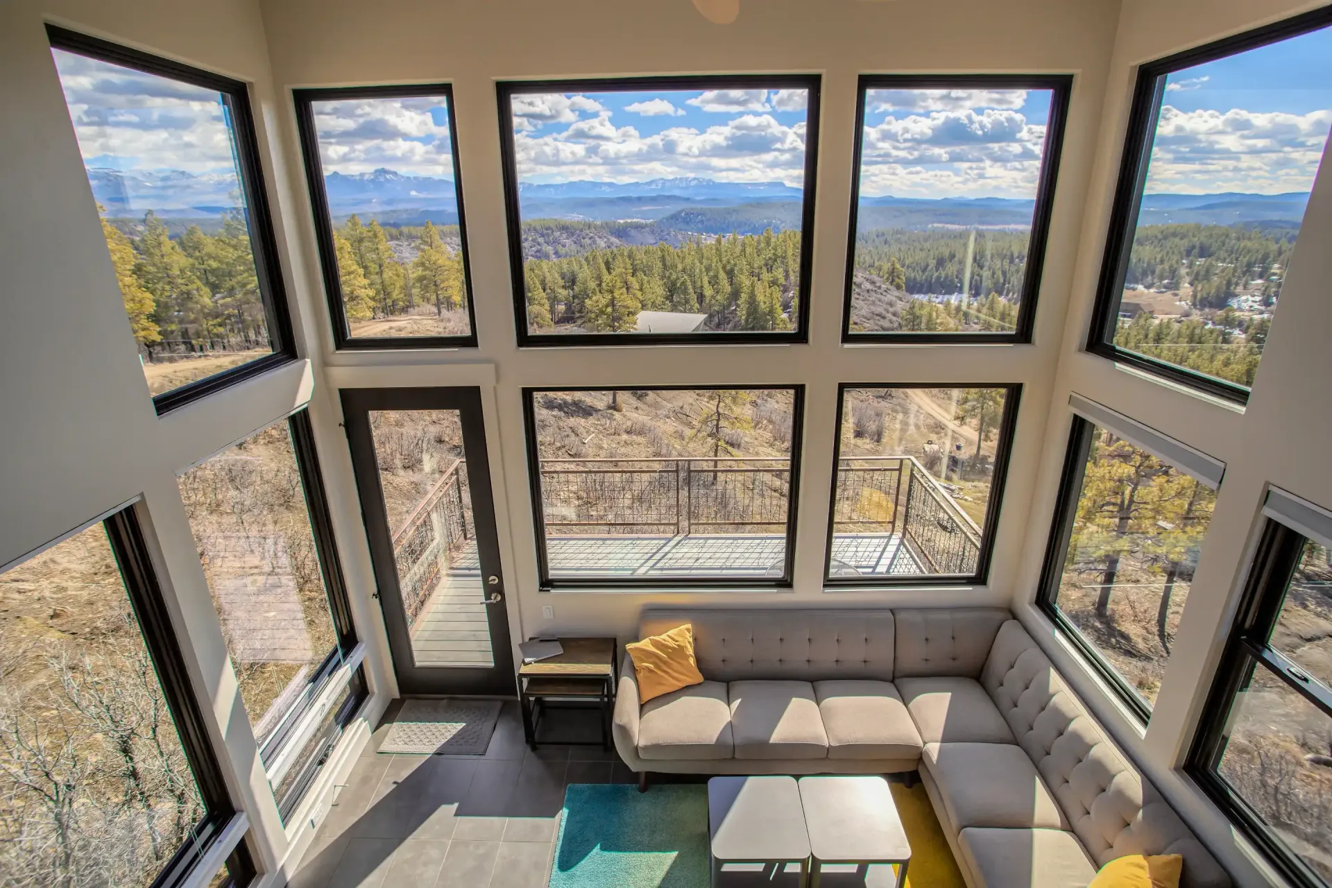 High-angle view of a sunlit living room with floor-to-ceiling windows overlooking a pine forest and distant mountains.