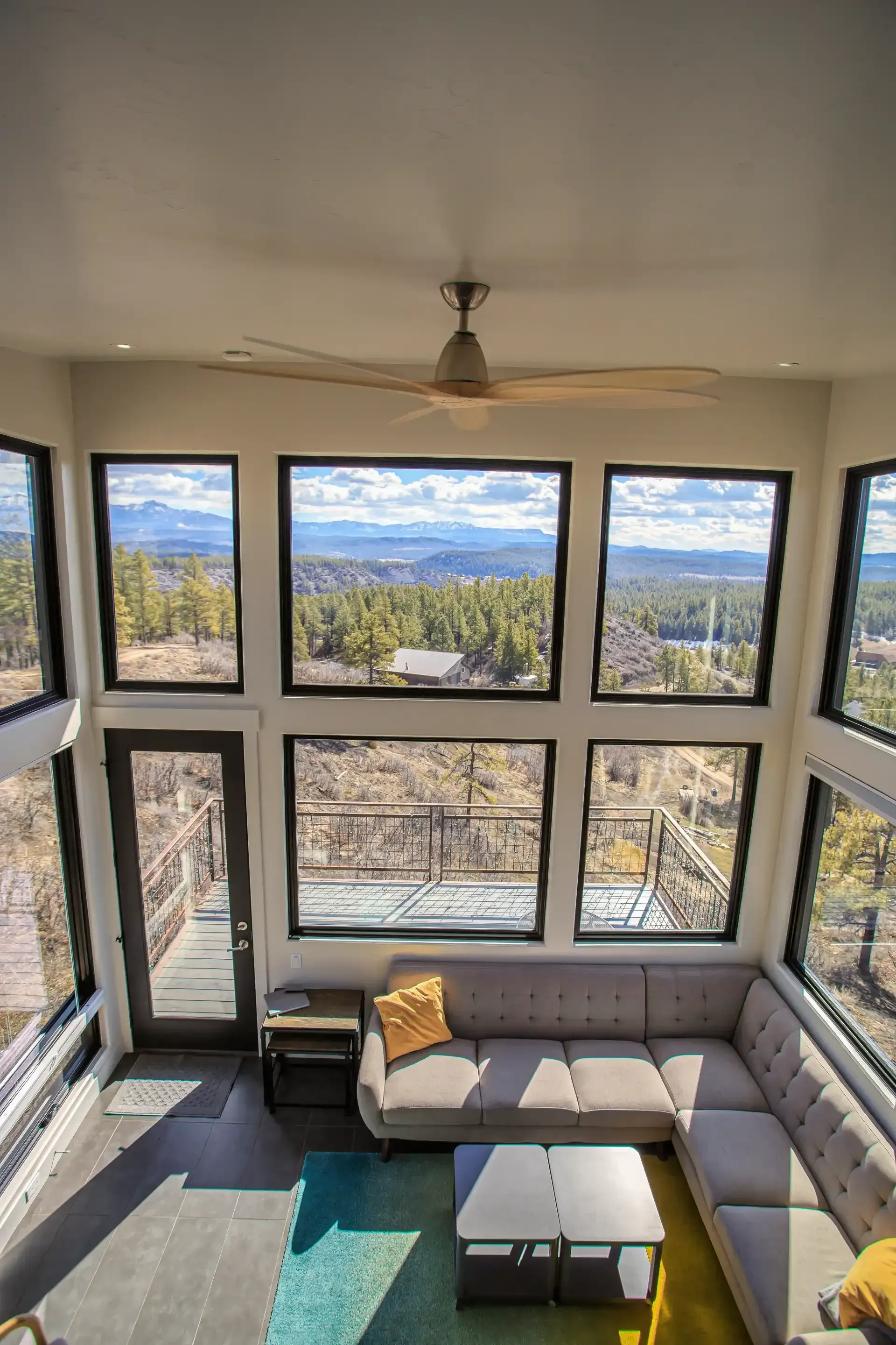 A cozy sunroom features a grey sectional sofa, teal and yellow rugs, and a ceiling fan, overlooking a forested mountain view.