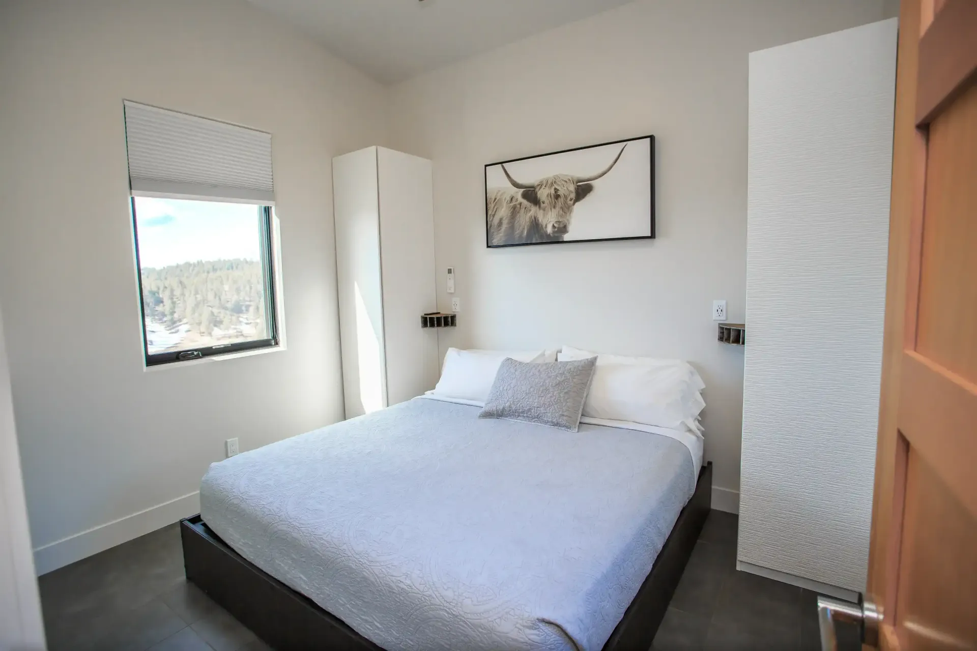 A minimalist bedroom featuring a queen bed with gray bedding, a tall white wardrobe, a window, and highland cow wall art.