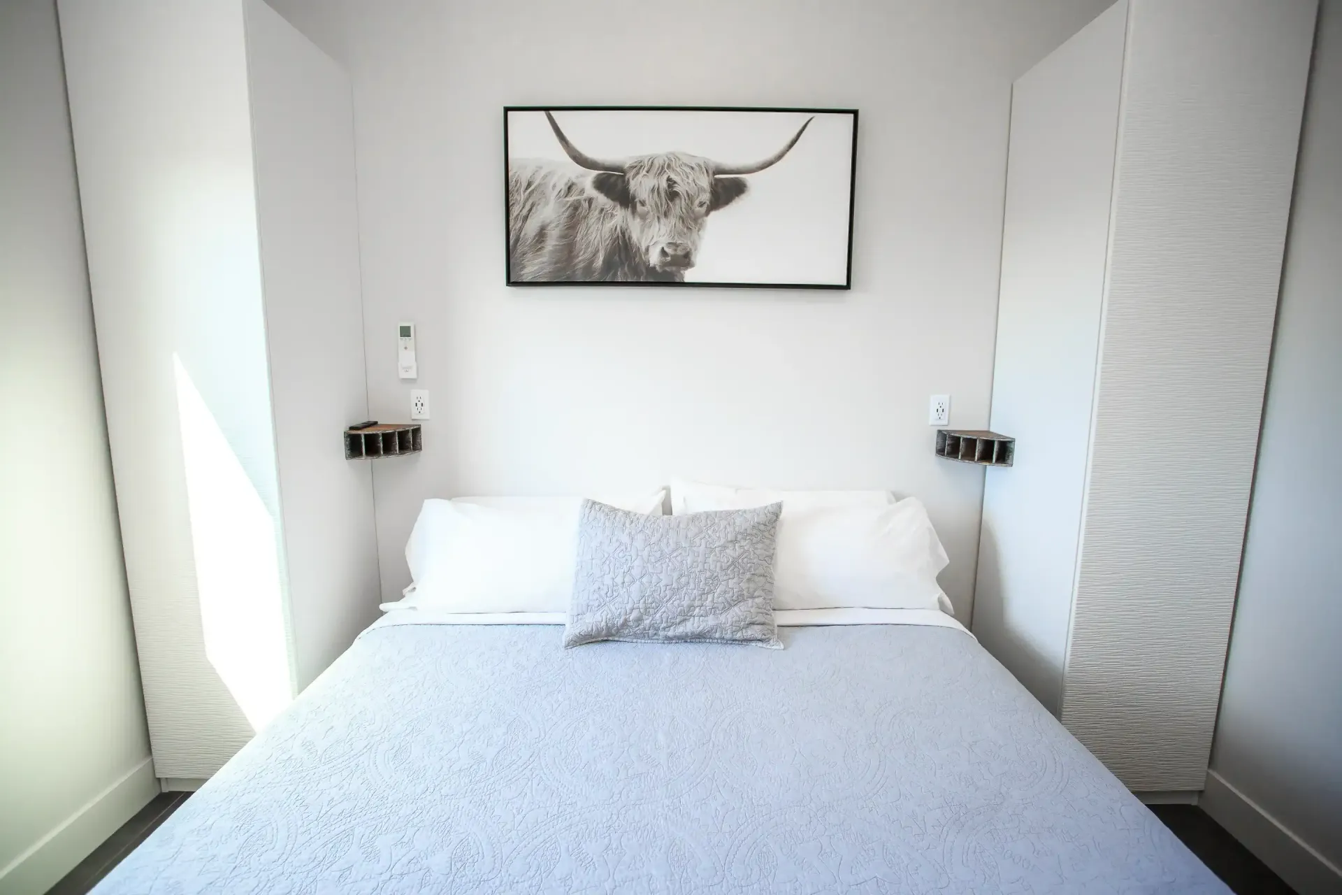 A bedroom with a bed featuring a light blue quilt and a Highland cow print hanging on the white wall between two closets.