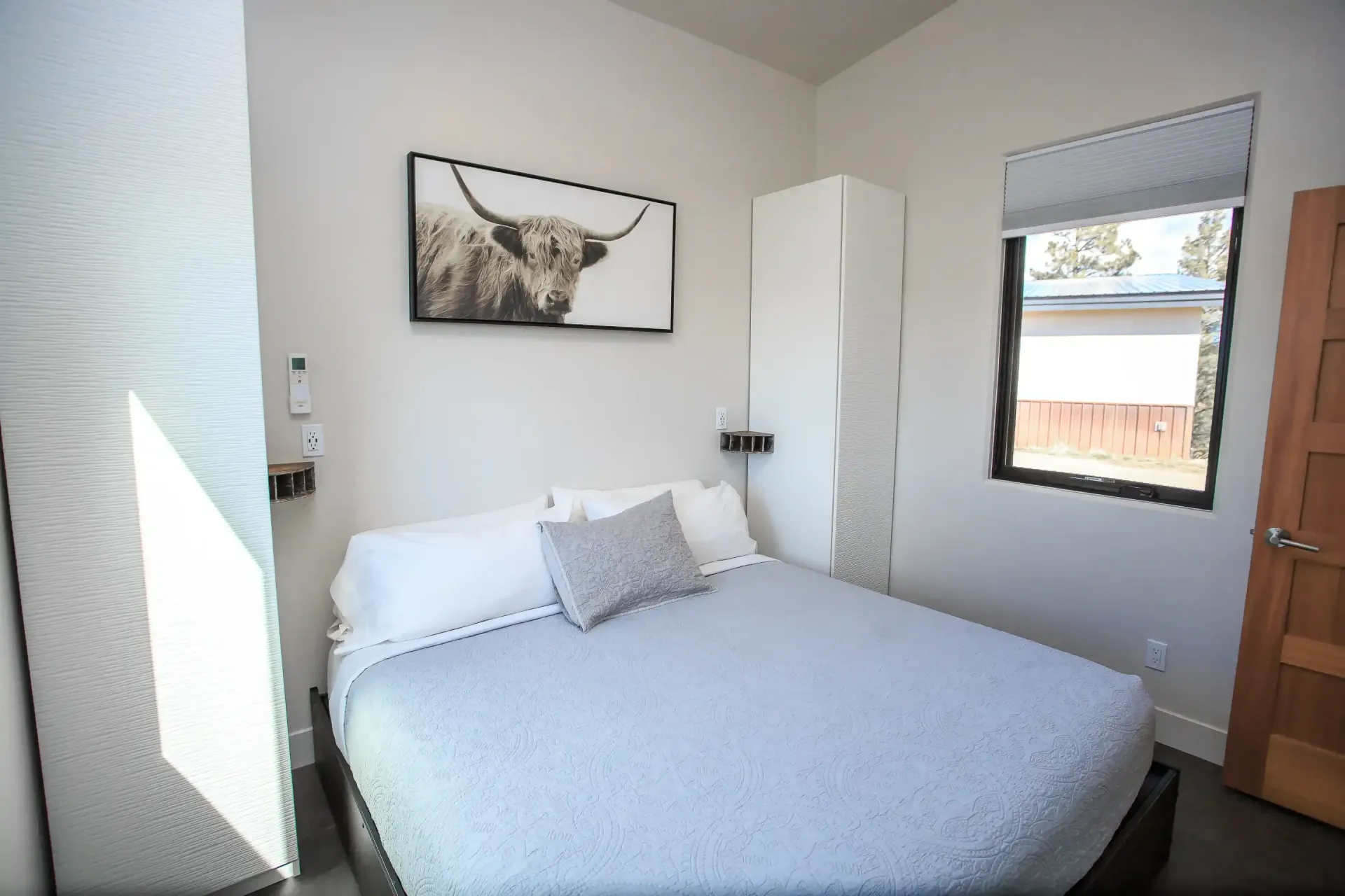 A small bedroom with a bed featuring a light blue quilt, a gray accent pillow, and a Highland cow print on the wall.