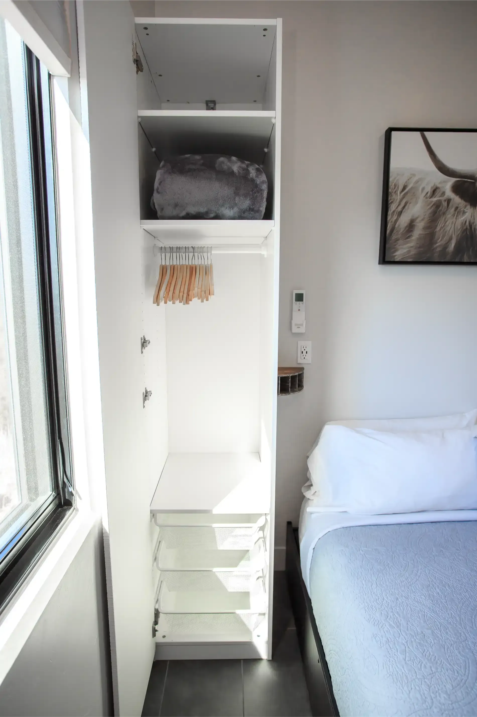 A white closet with shelves, a hanging rod, and drawers beside a bed in a room with a window and a wall art piece.
