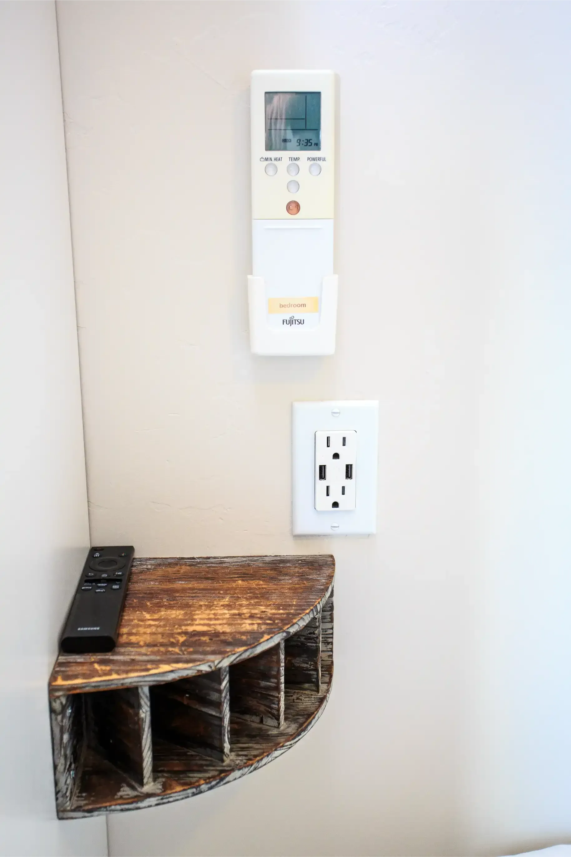 A wall-mounted corner shelf with a remote, an air conditioner remote in a holder, and an electrical outlet below it.