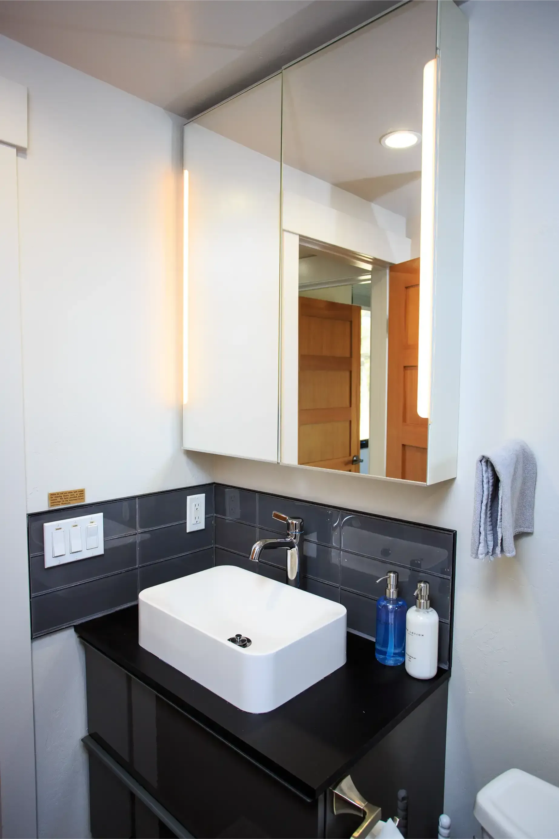 A modern bathroom sink with a white rectangular basin, black vanity, gray tiled backsplash, and an illuminated mirror.
