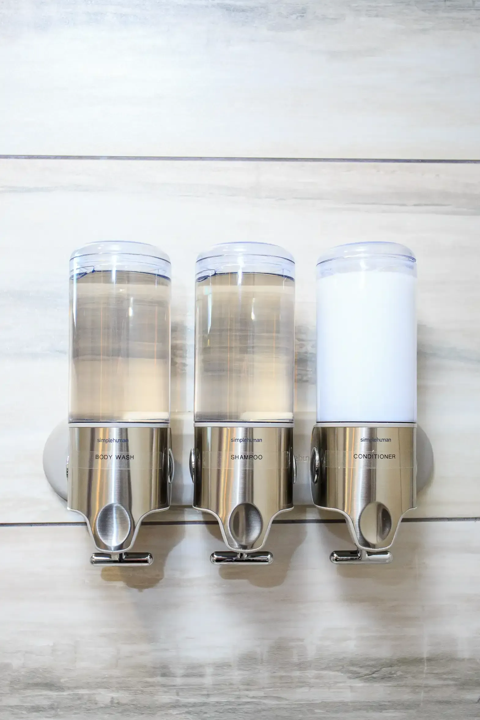 Wall-mounted soap dispenser with three containers: two clear, one filled with white opaque liquid, on a marble-tiled wall.