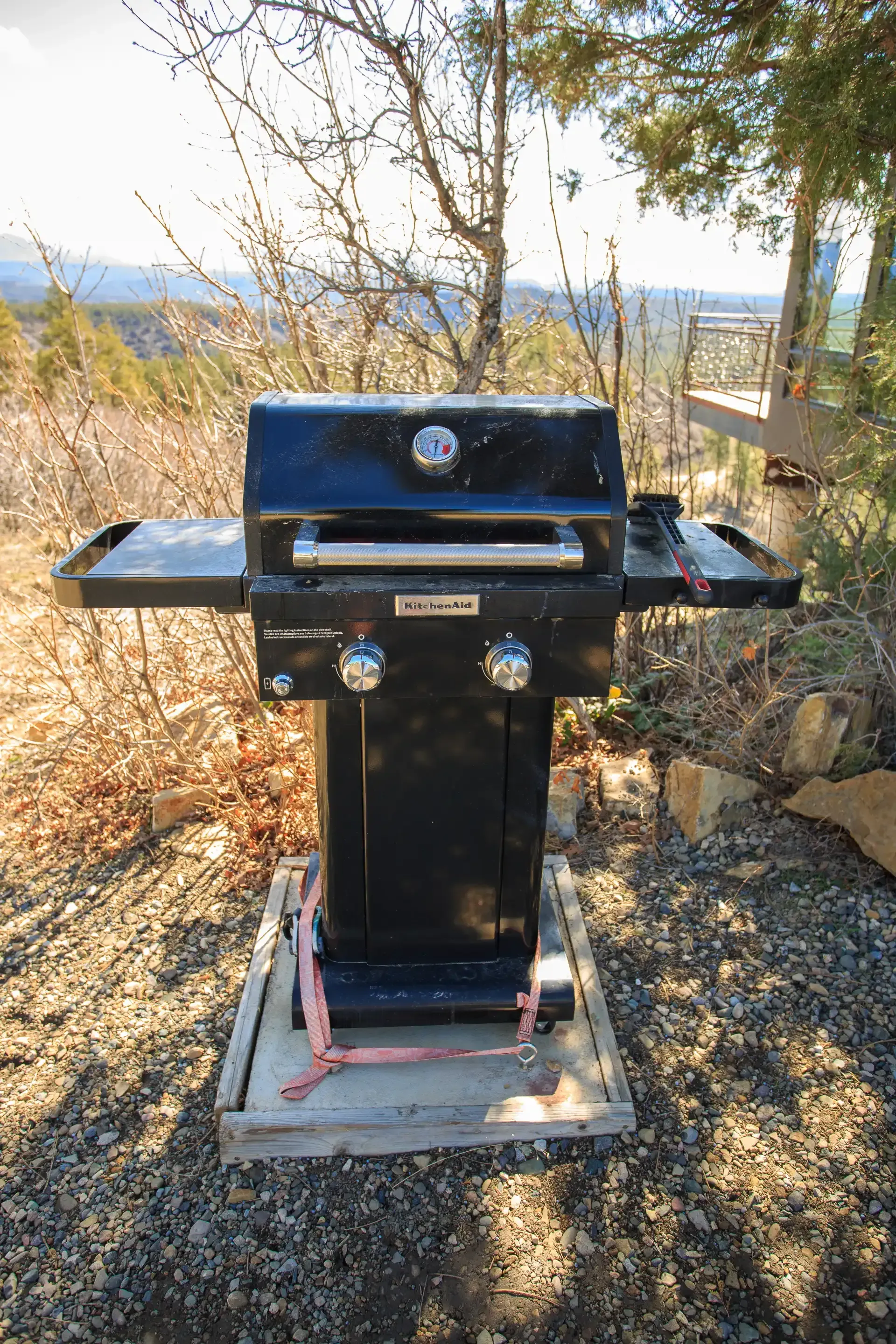 A black two-burner gas grill secured to a wooden base on a gravel surface, with trees and a mountainous view behind it.