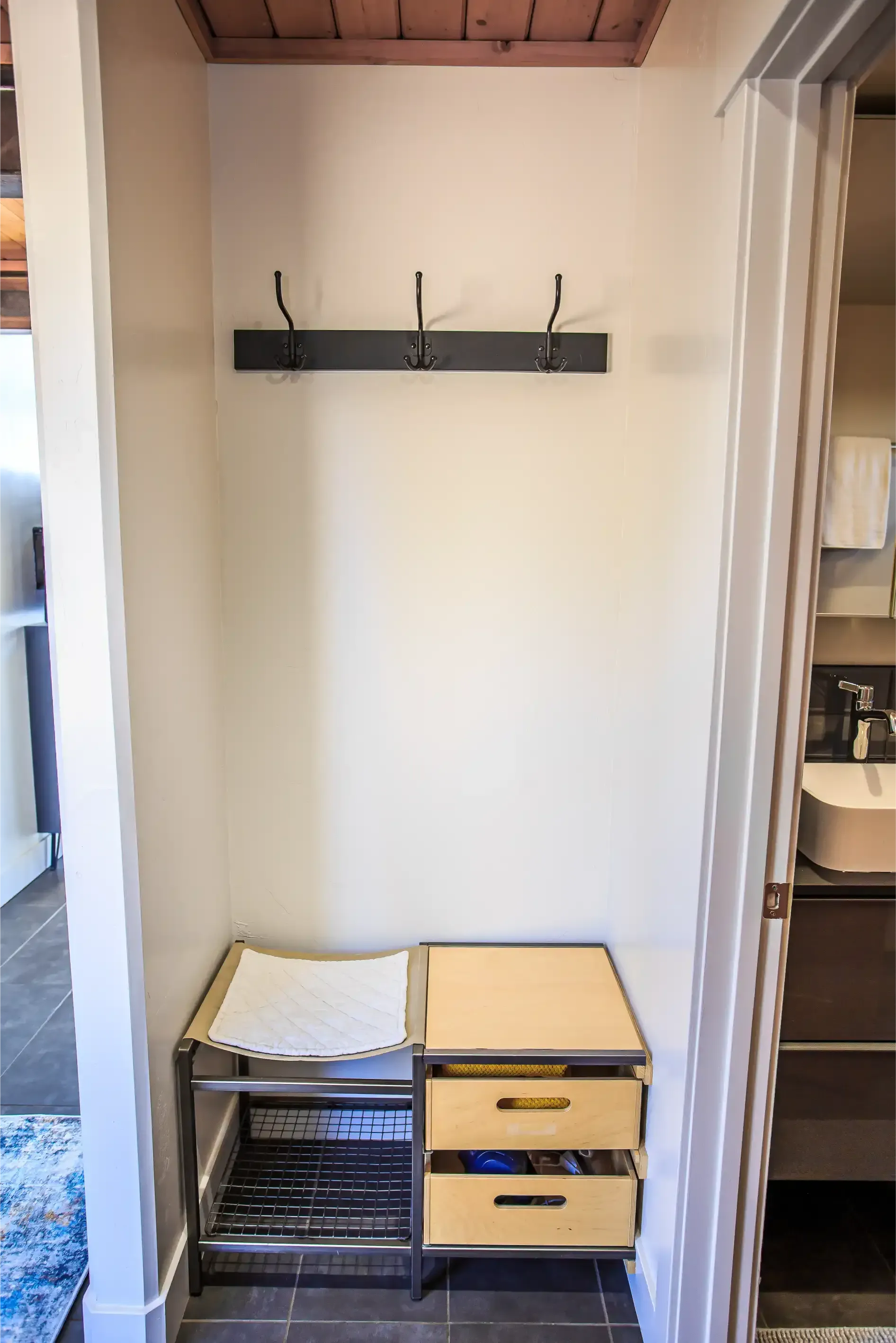 An entryway nook with a wall-mounted coat rack featuring three metal hooks above a two-section metal bench with drawers.