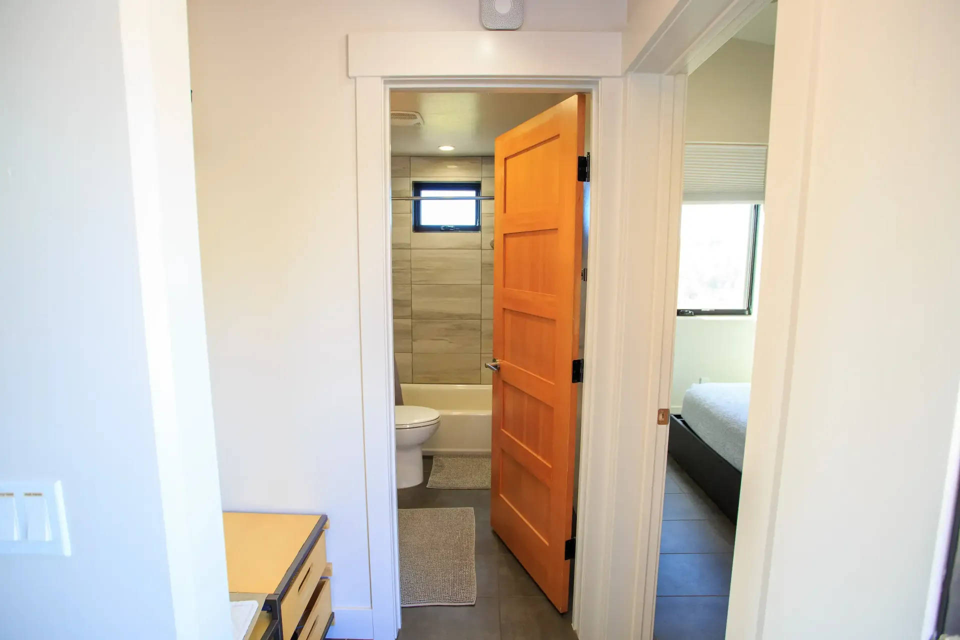 A view from a hallway into a bathroom with a light wood door, tiled floors, and a glimpse of a bedroom to the right.