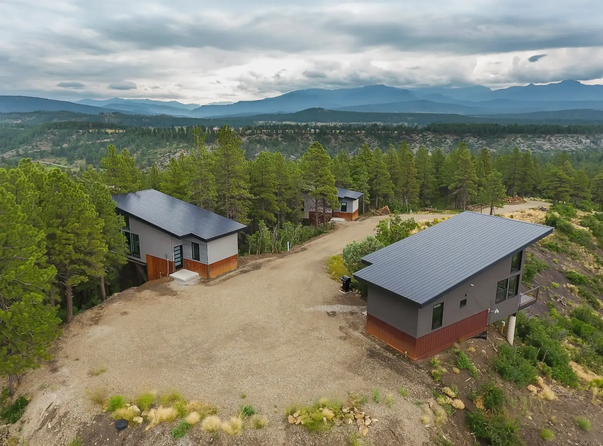 Two modern houses with metal roofs on a rocky, tree-lined ridge overlooking a scenic valley under a cloudy sky.