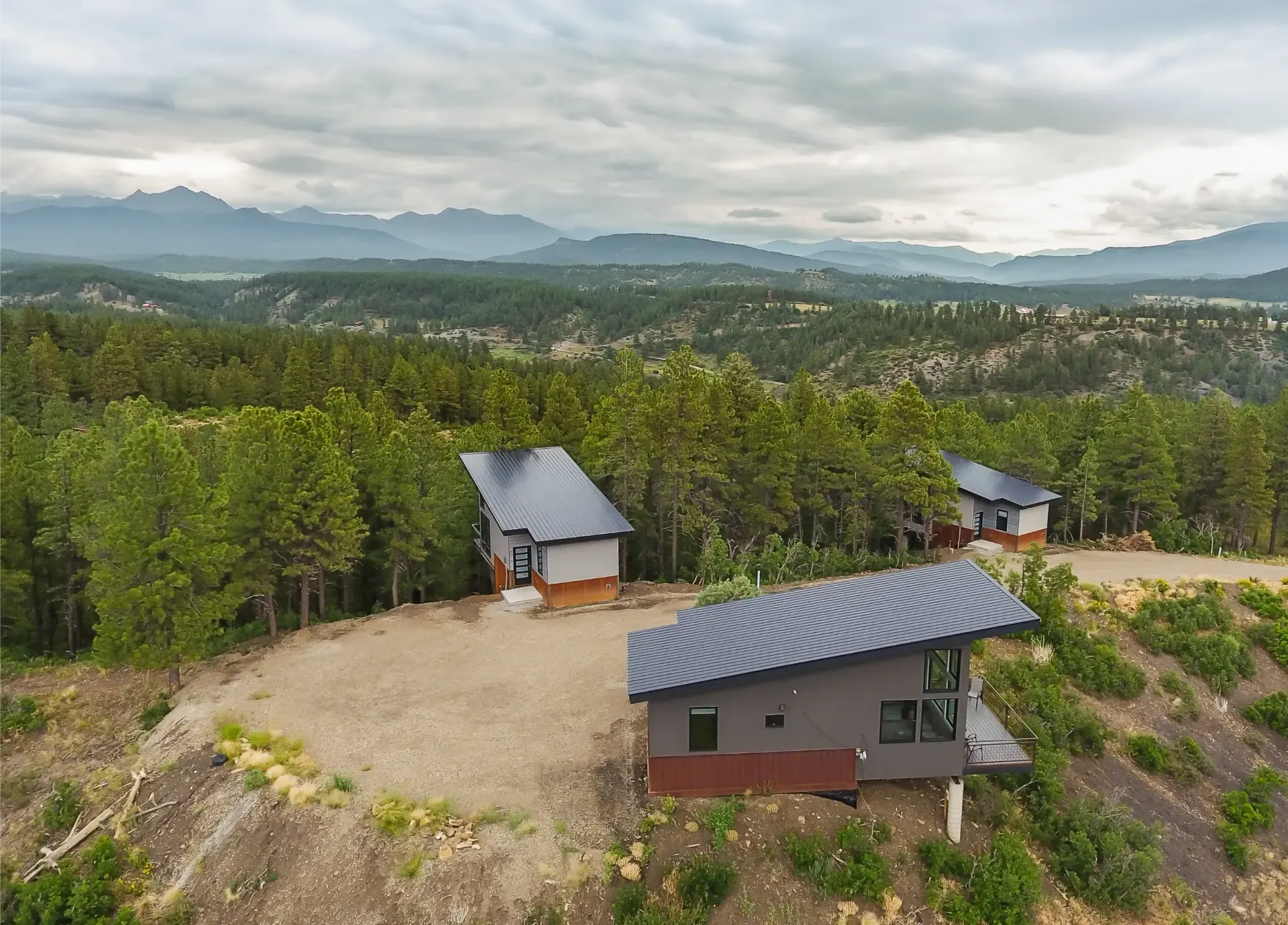 Two modern, grey buildings with slanted roofs sit on a hillside overlooking a pine forest and distant mountain range.