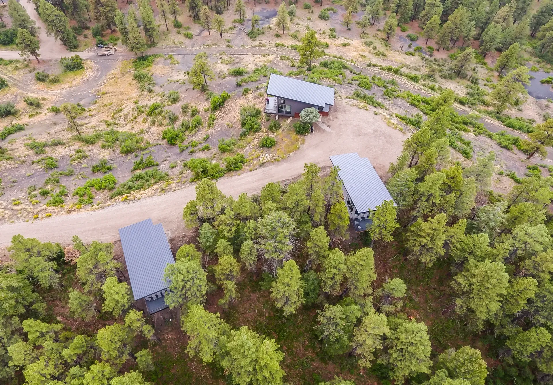 An aerial view of three small, modern cabins with dark roofs nestled within a sparse pine forest next to a dirt road.