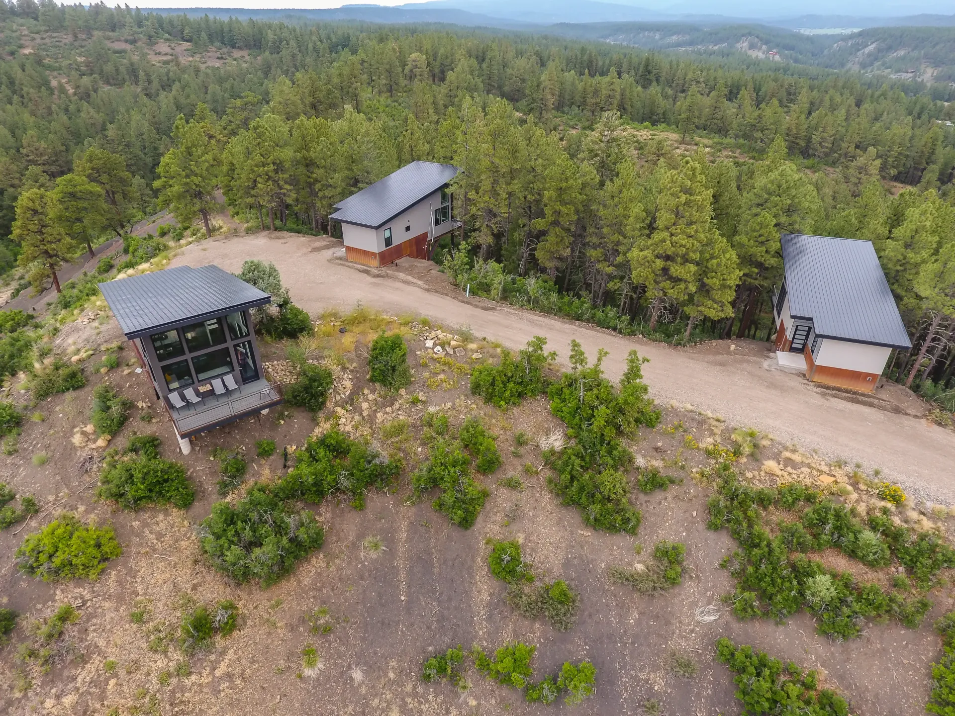 Aerial view of three modern, multi-level cabins with dark roofs tucked into a forested hillside along a dirt path.