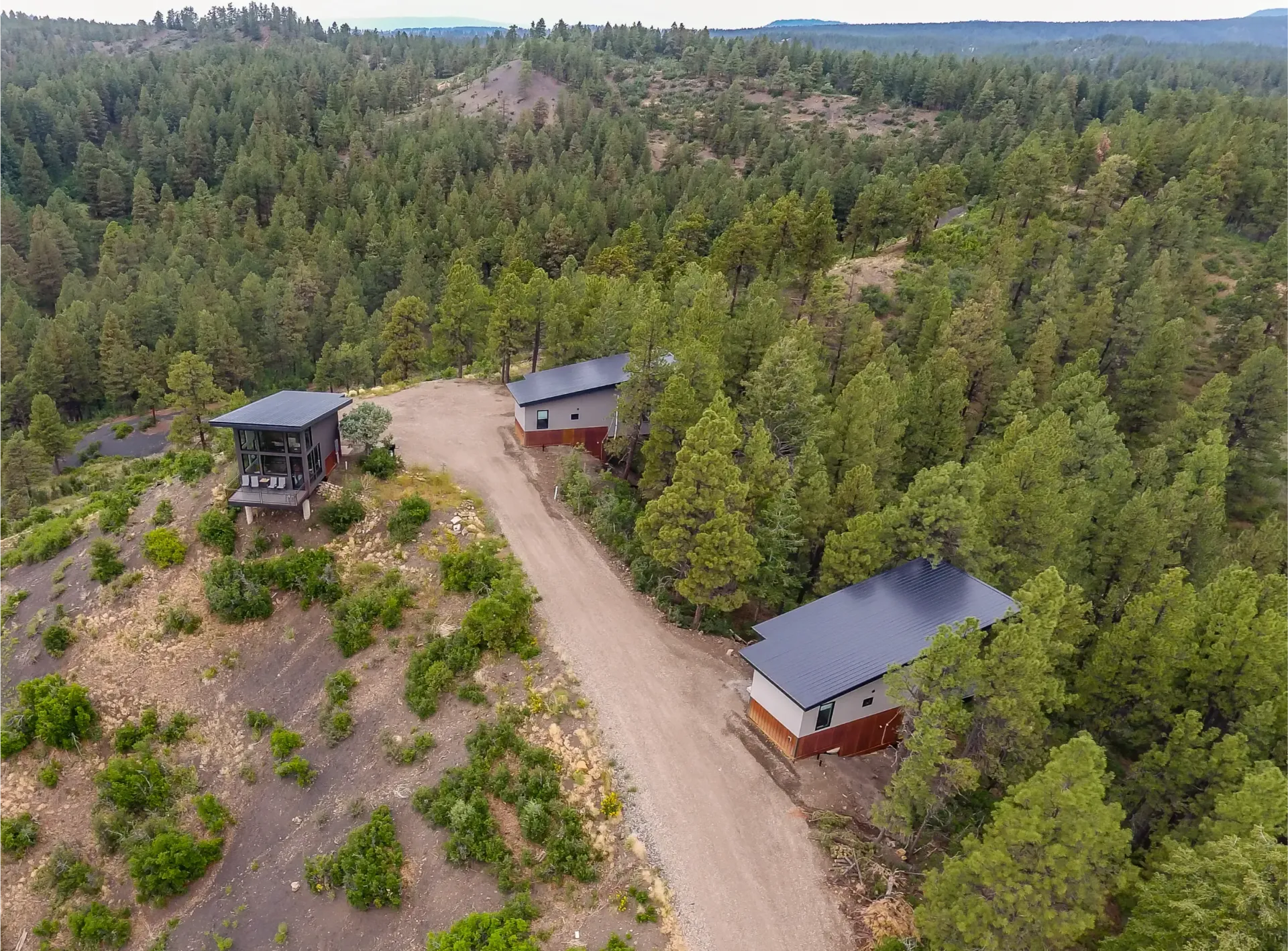An aerial view of three modern cabins with dark roofs nestled along a dirt road in a dense, green pine forest.