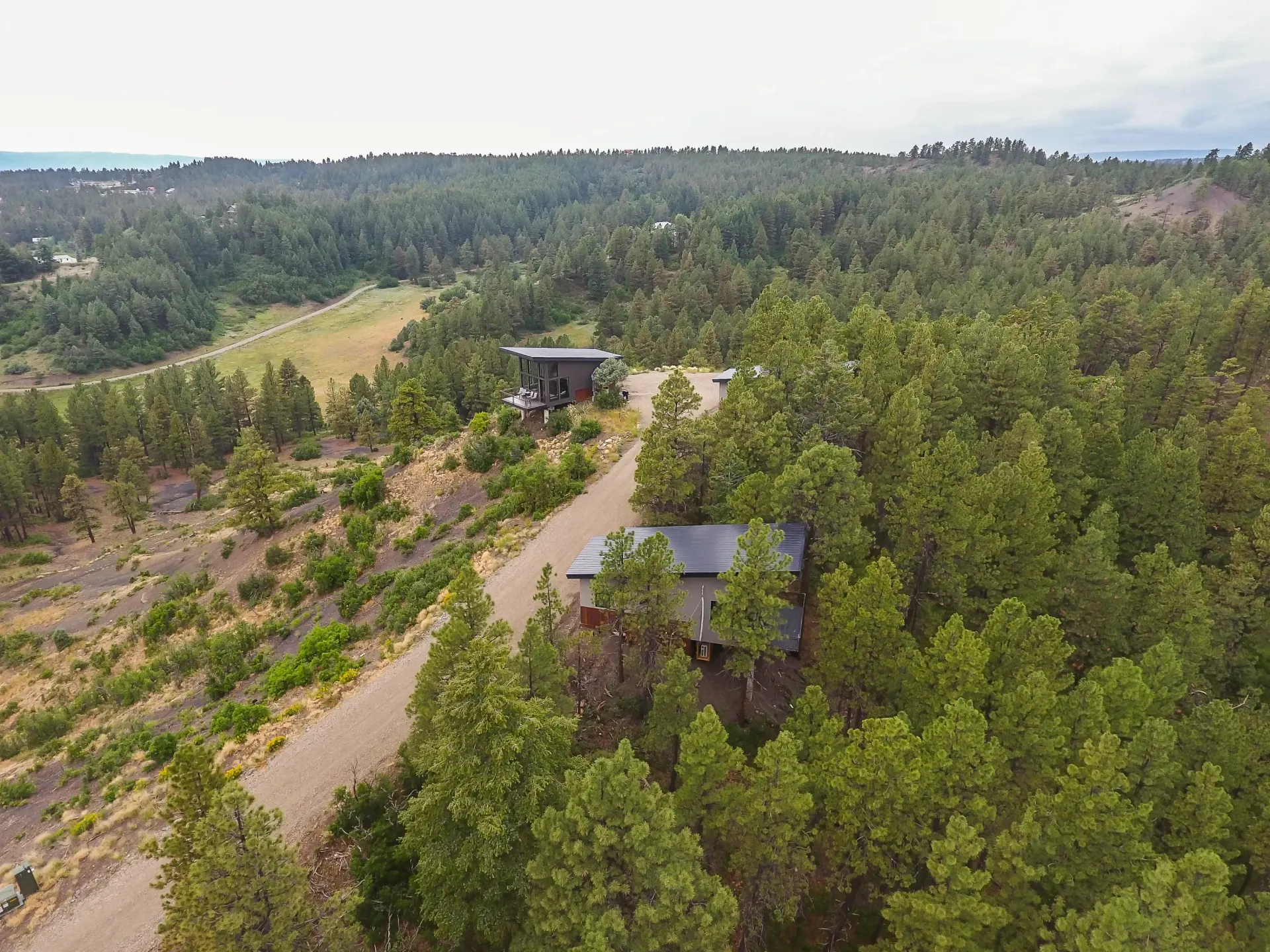 An aerial view of two modern homes nestled within a dense, hilly evergreen forest alongside a gravel driveway.