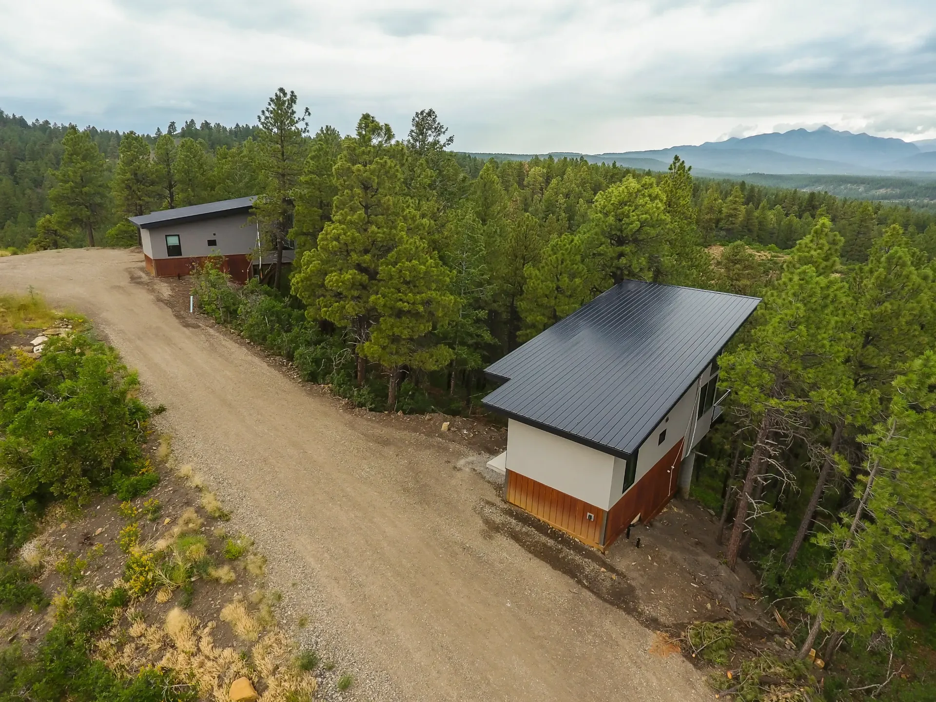Two modern cabins with dark, sloped roofs sit on a gravel road in a forested landscape with distant mountains.