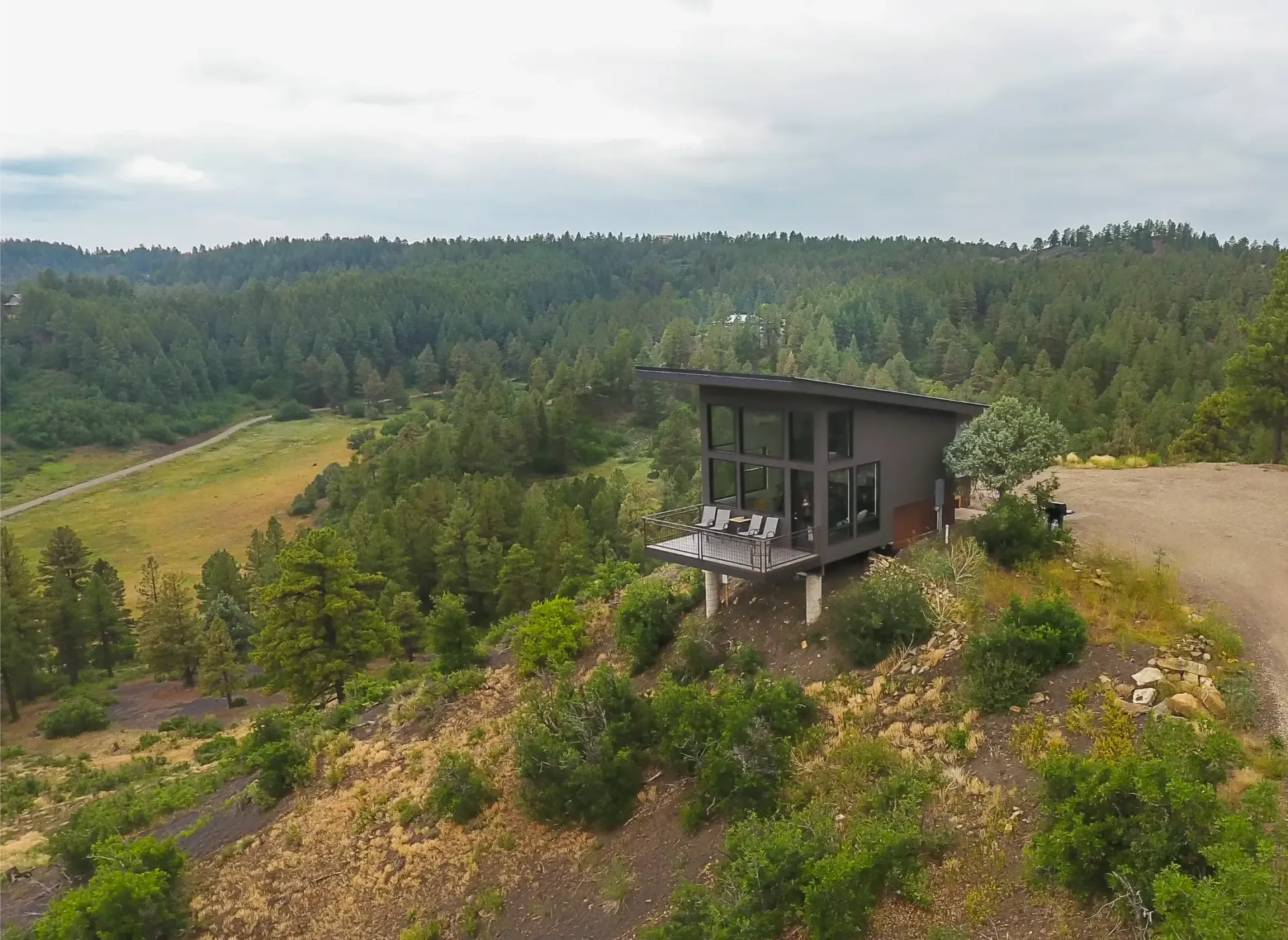 A modern, dark-toned cabin perched on a hillside overlooking a vast forest and valley.