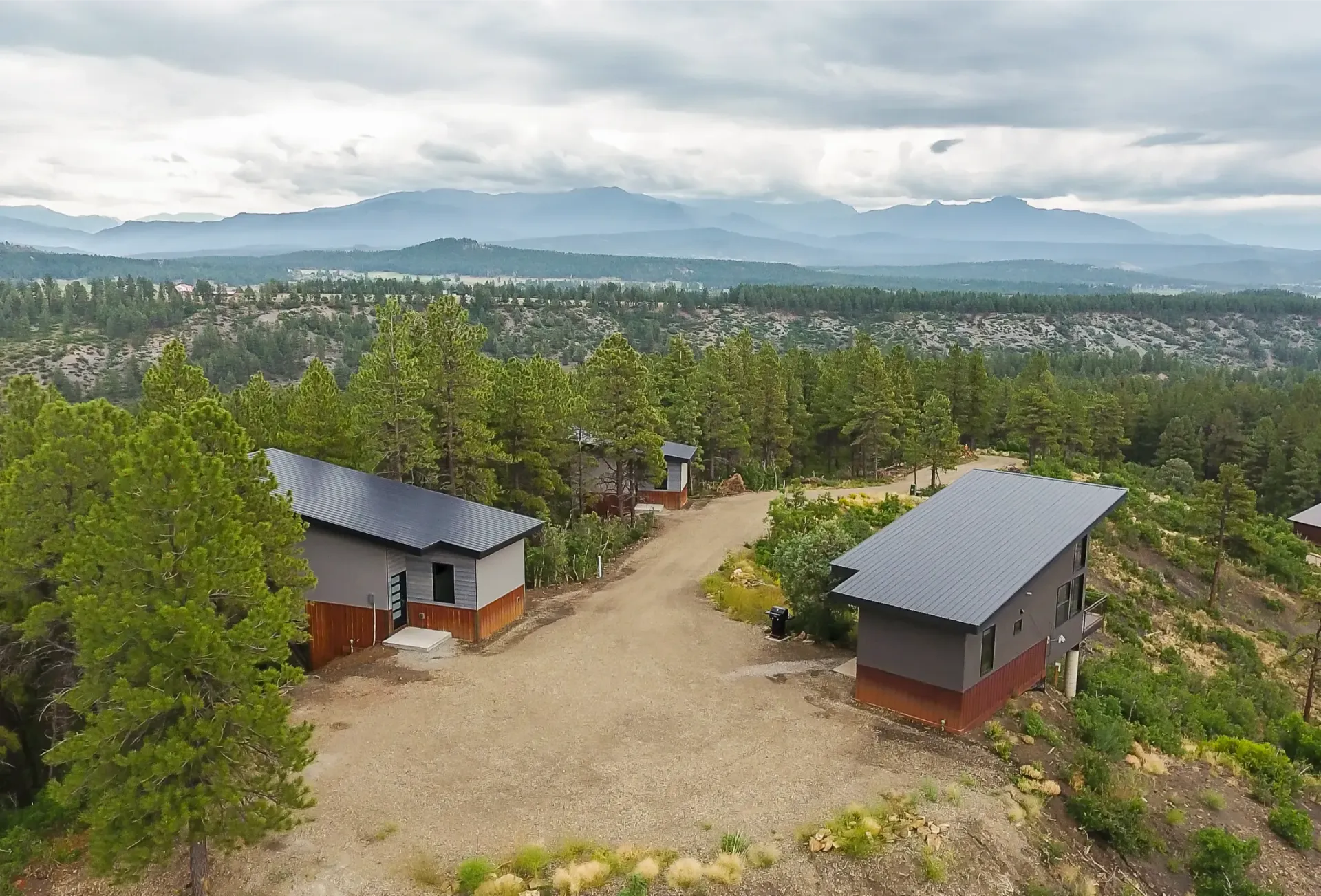 Two modern houses with dark slanted roofs sit on a gravel path surrounded by trees, with mountains in the background.