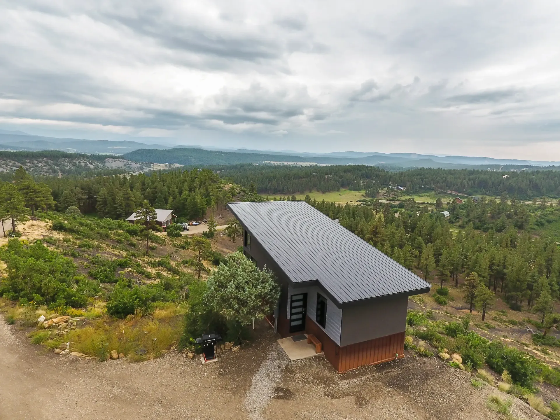 An aerial view of a modern, flat-roofed cabin perched on a hillside overlooking a vast forest and valley under a cloudy sky.
