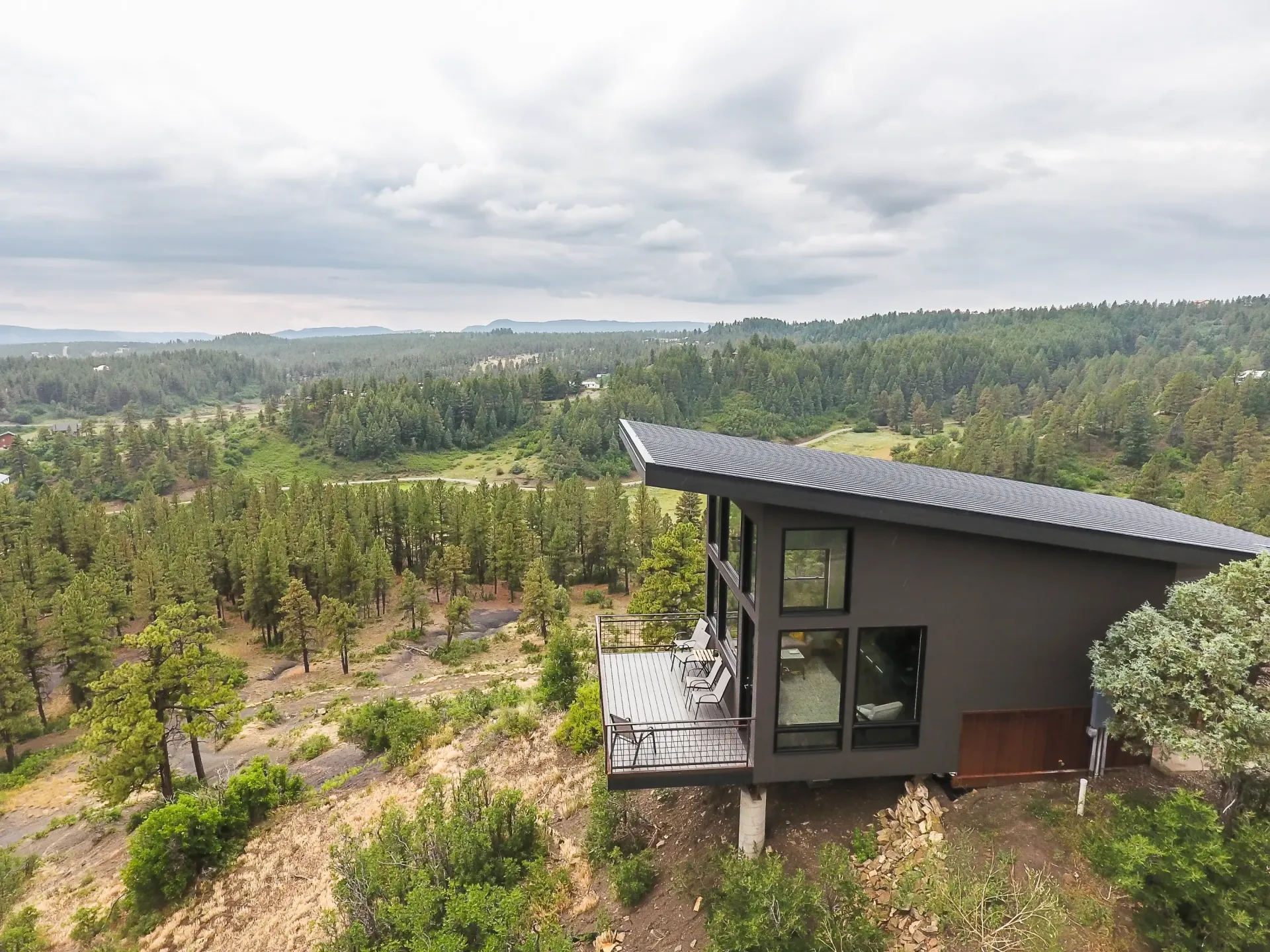 A modern dark gray house with a slanted roof sits on a hillside overlooking a vast, wooded valley under a cloudy sky.
