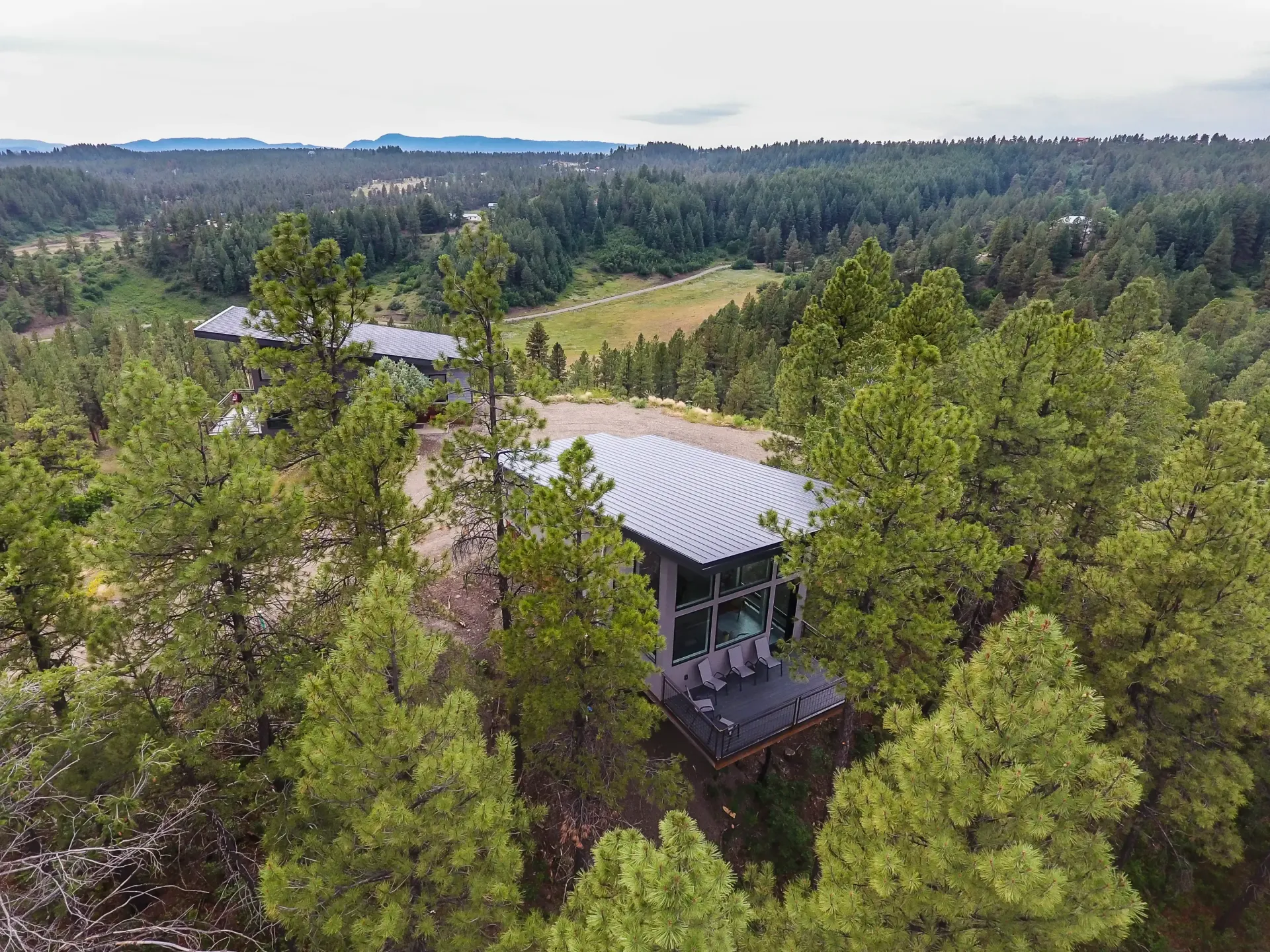 Aerial view of two modern, flat-roofed houses nestled among dense green pine trees in a hilly, forest landscape.