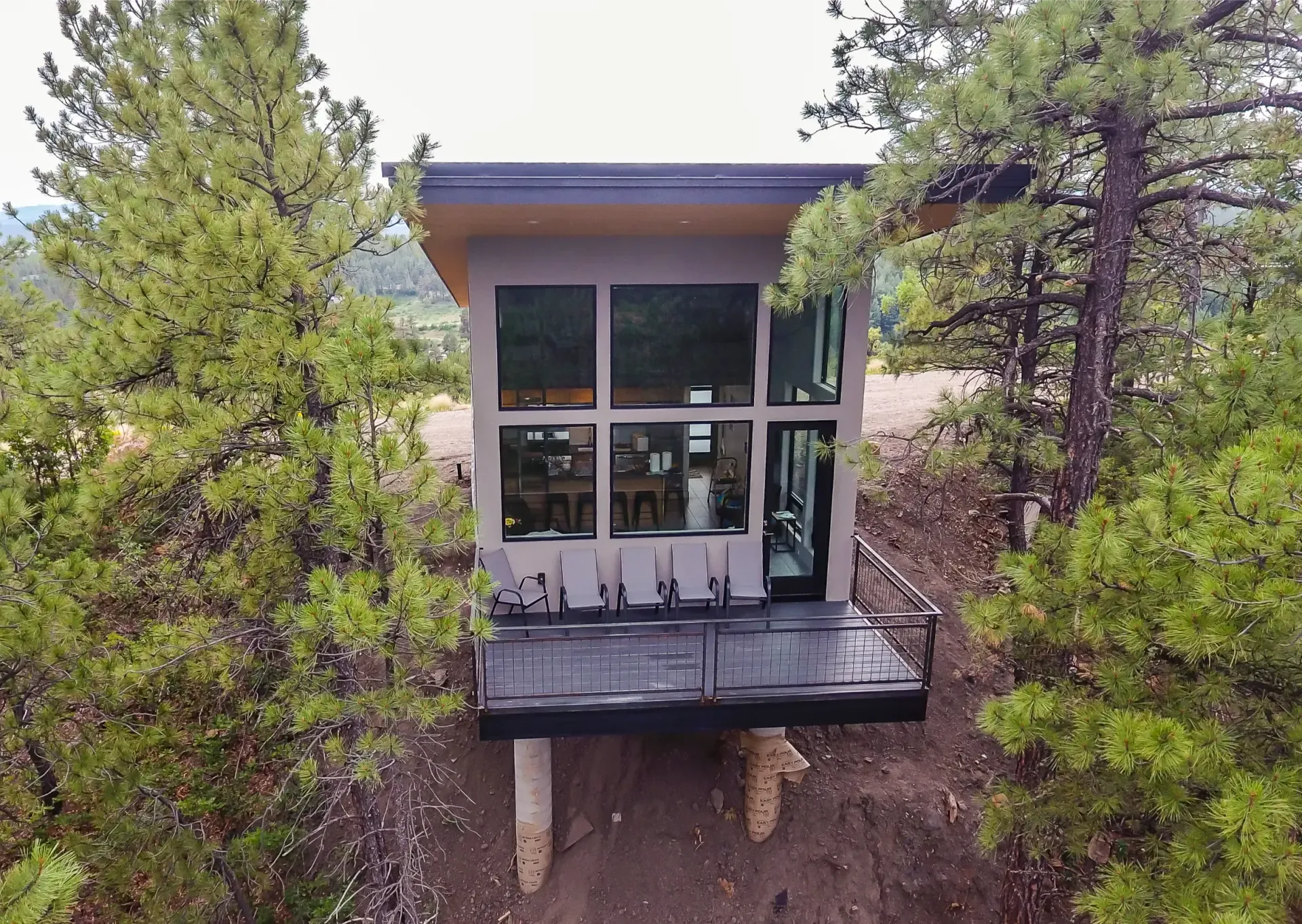 A modern, boxy cabin on stilts nestled in a pine forest, featuring large glass windows and a dark, low-profile deck.