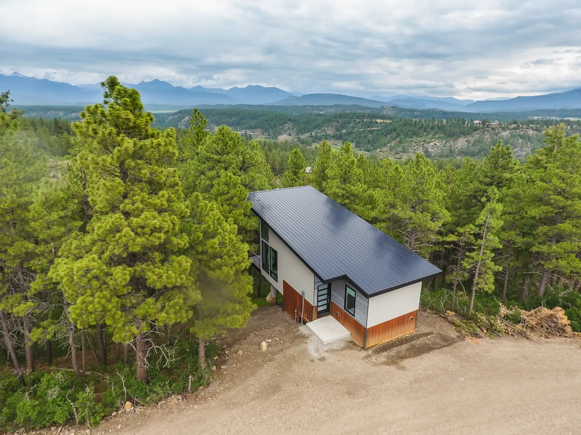 A modern cabin with a dark corrugated roof sits nestled among pine trees on a hillside overlooking distant mountains.