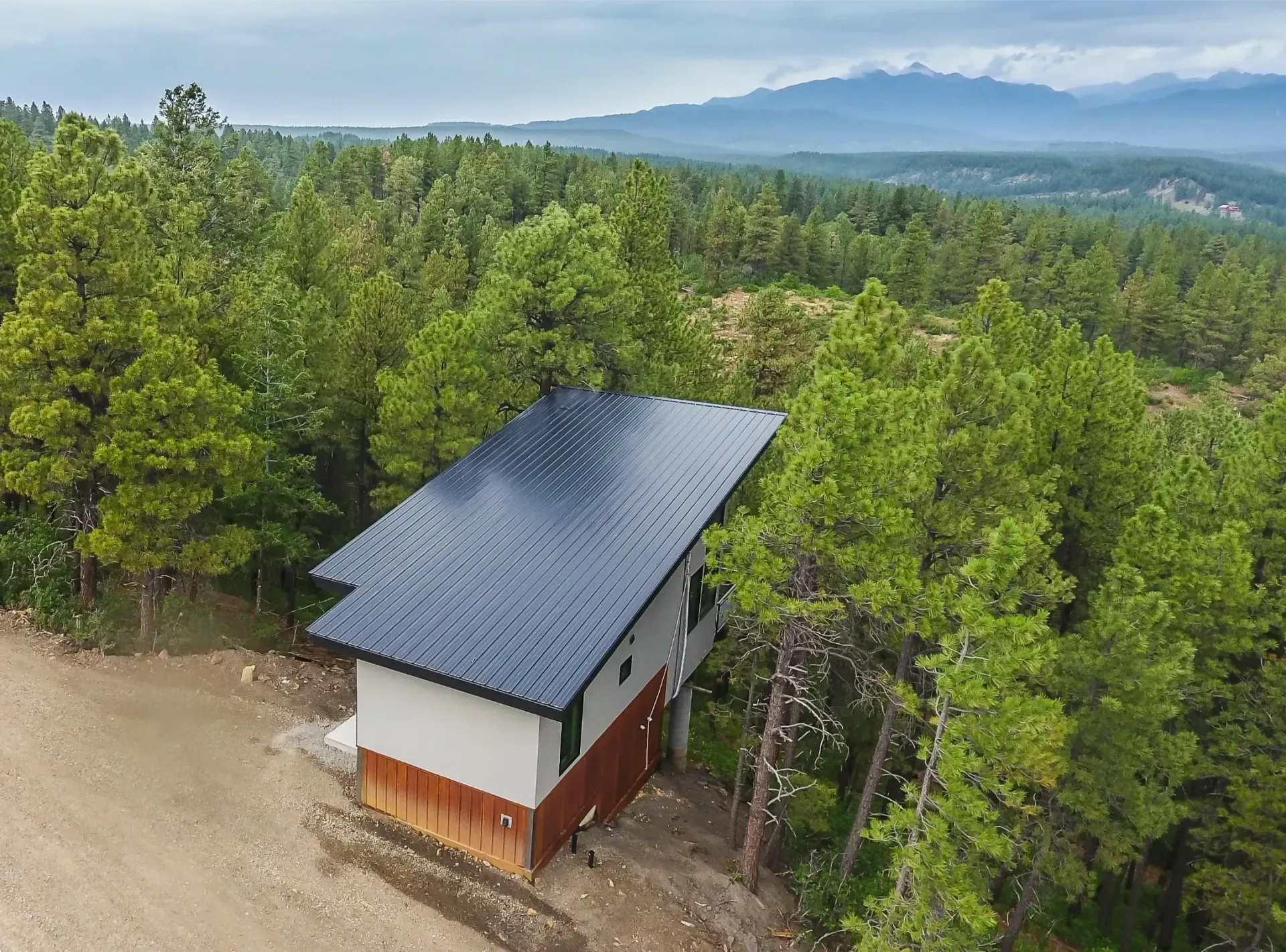 An aerial view of a modern two-story house with a rippled dark roof, nestled in a dense pine forest near mountains.