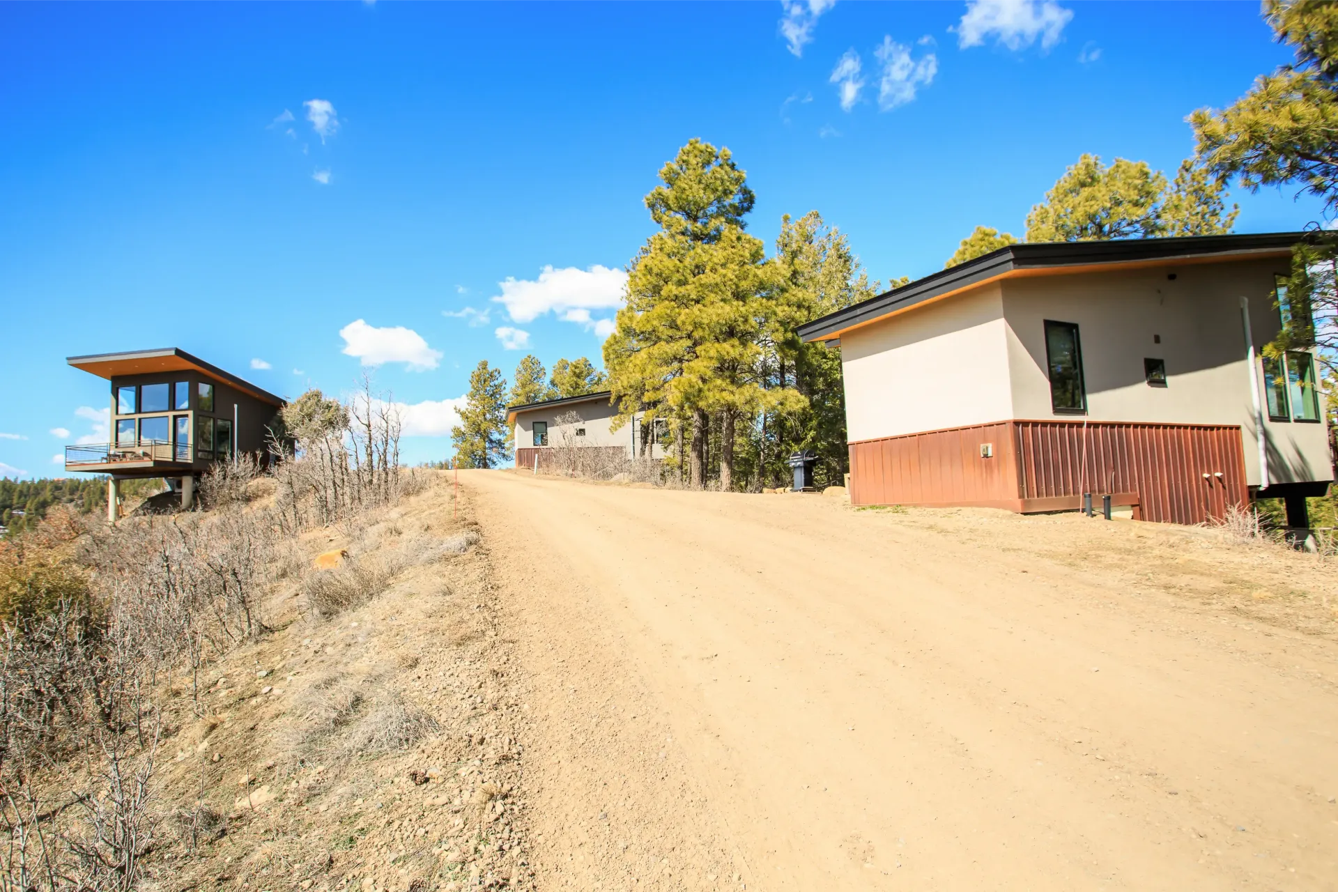 Two modern cabins with slanted roofs sit on a hillside alongside a dirt path under a clear blue sky.