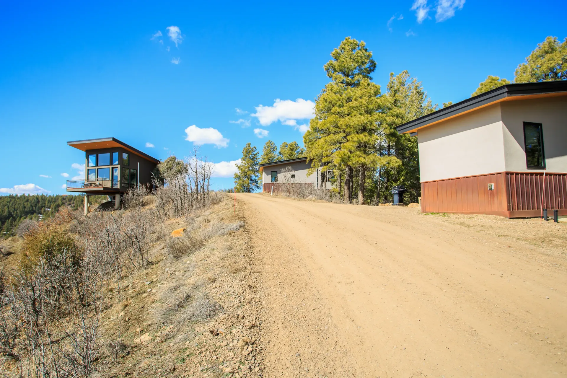 A dirt path leads uphill toward a modern, flat-roofed house and a small, glass-walled structure under a clear blue sky.