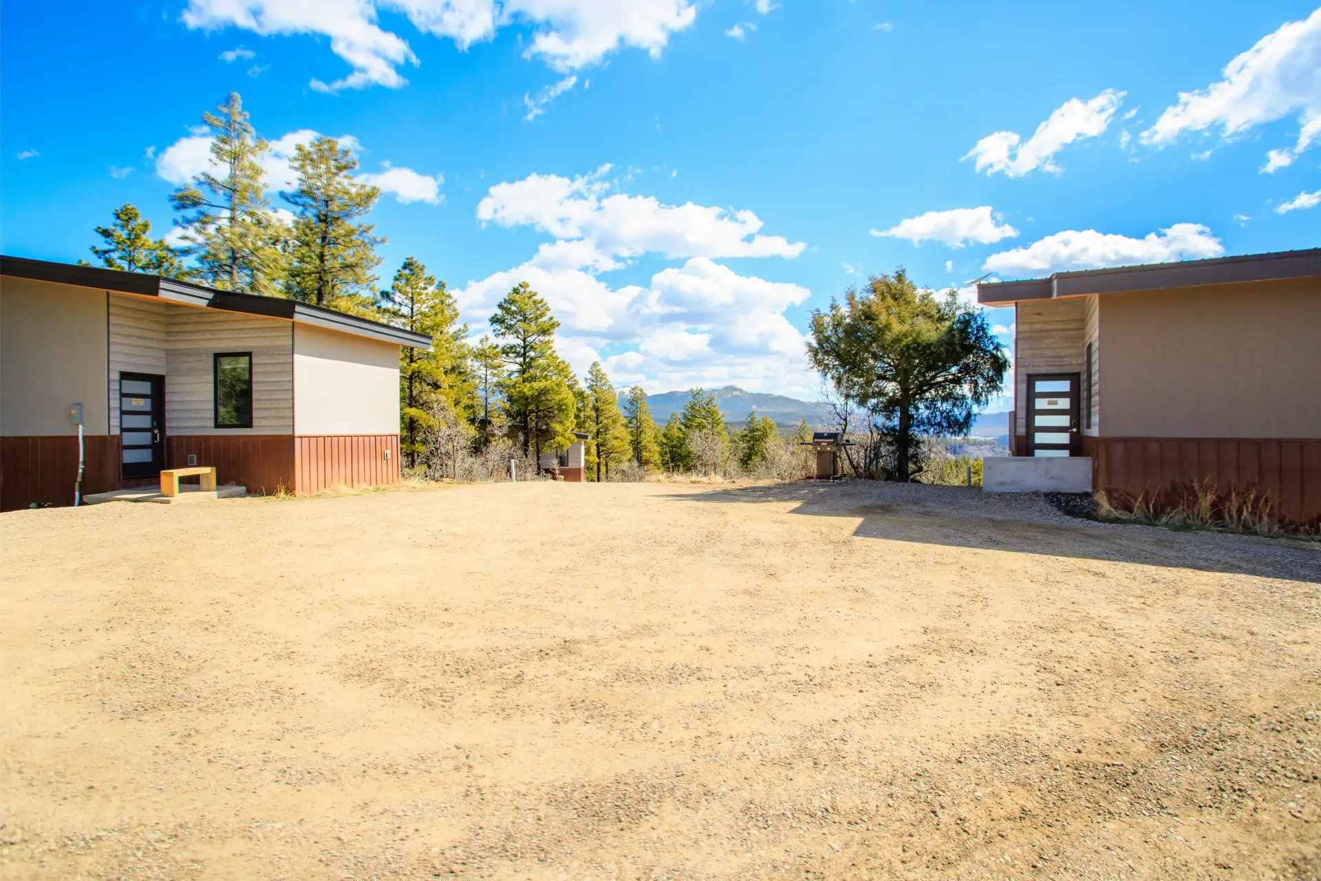 Two modern structures face each other across a dry, grassy clearing with a background of pine trees and blue sky.