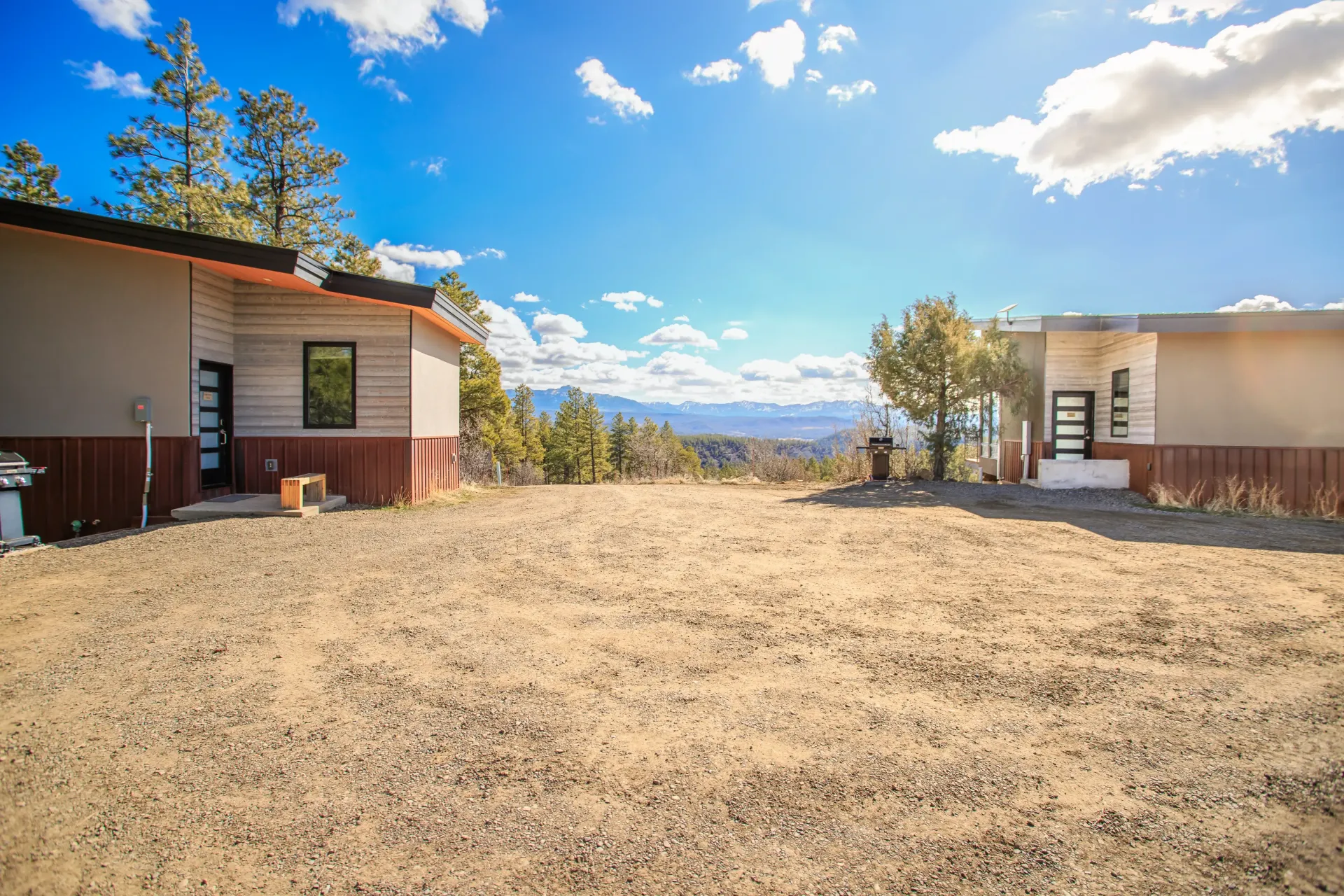 Two modern, light-colored houses separated by a dry, grass-covered yard under a blue sky with scattered white clouds.