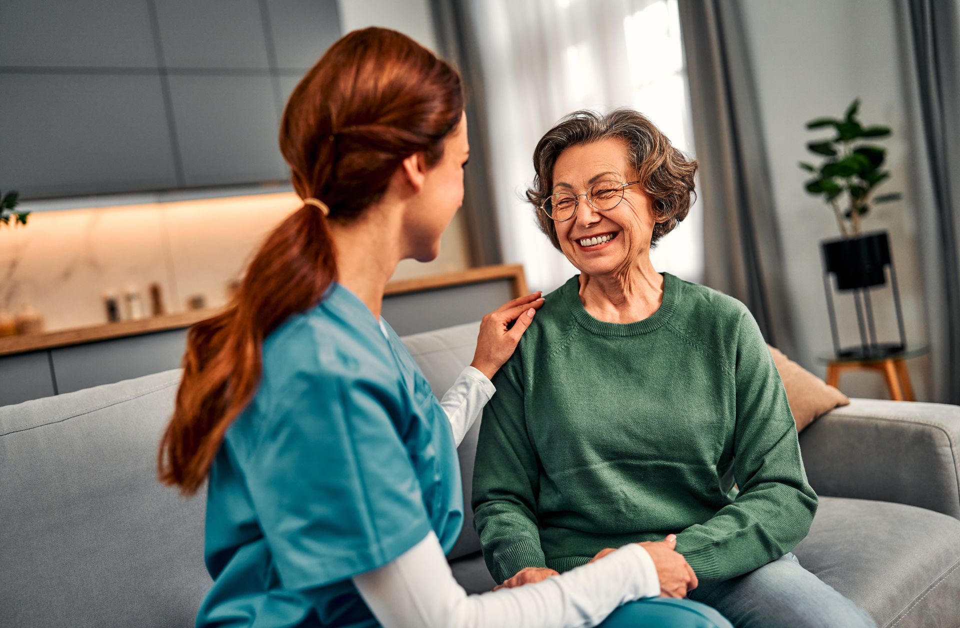 Caregiver speaking with a smiling patient on a couch in a bright living room