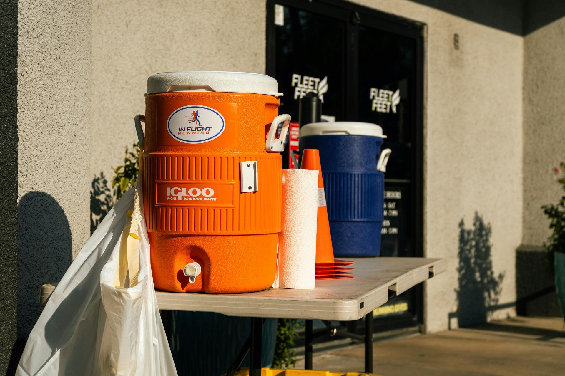 Orange and blue water coolers with cups on a table outside a building.