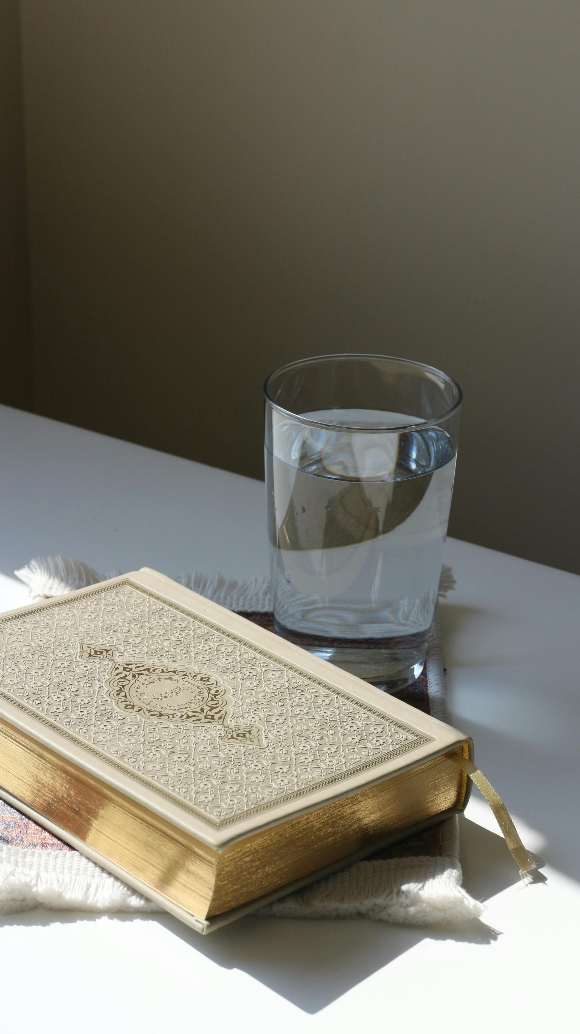 Book with gold-edged pages, glass of water, sunlight on a white surface.