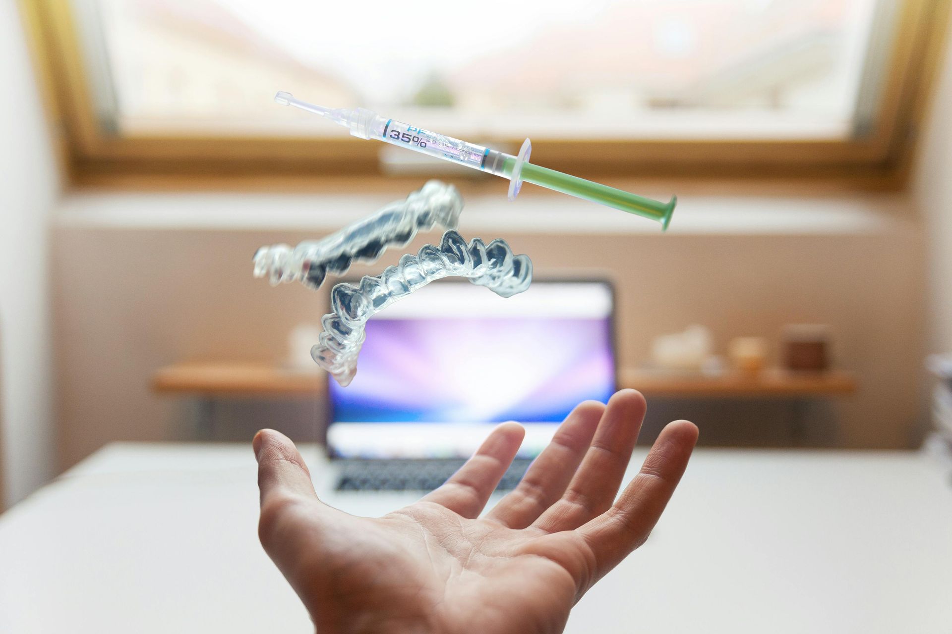 A hand is holding a syringe and dental braces in front of a laptop.
