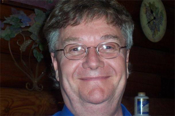 Man with glasses smiling, short brown hair, blue shirt, indoor setting.