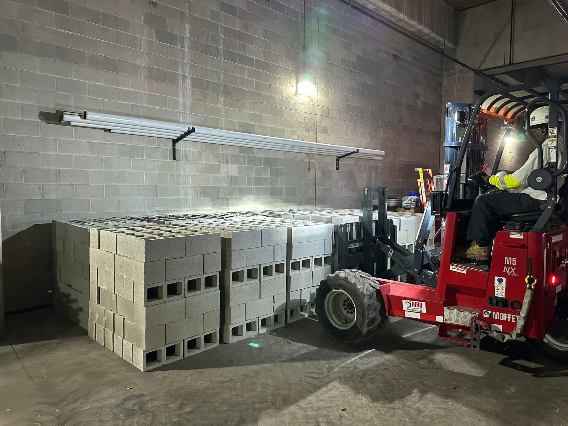 Forklift operator loading materials in a warehouse with stacked concrete blocks.