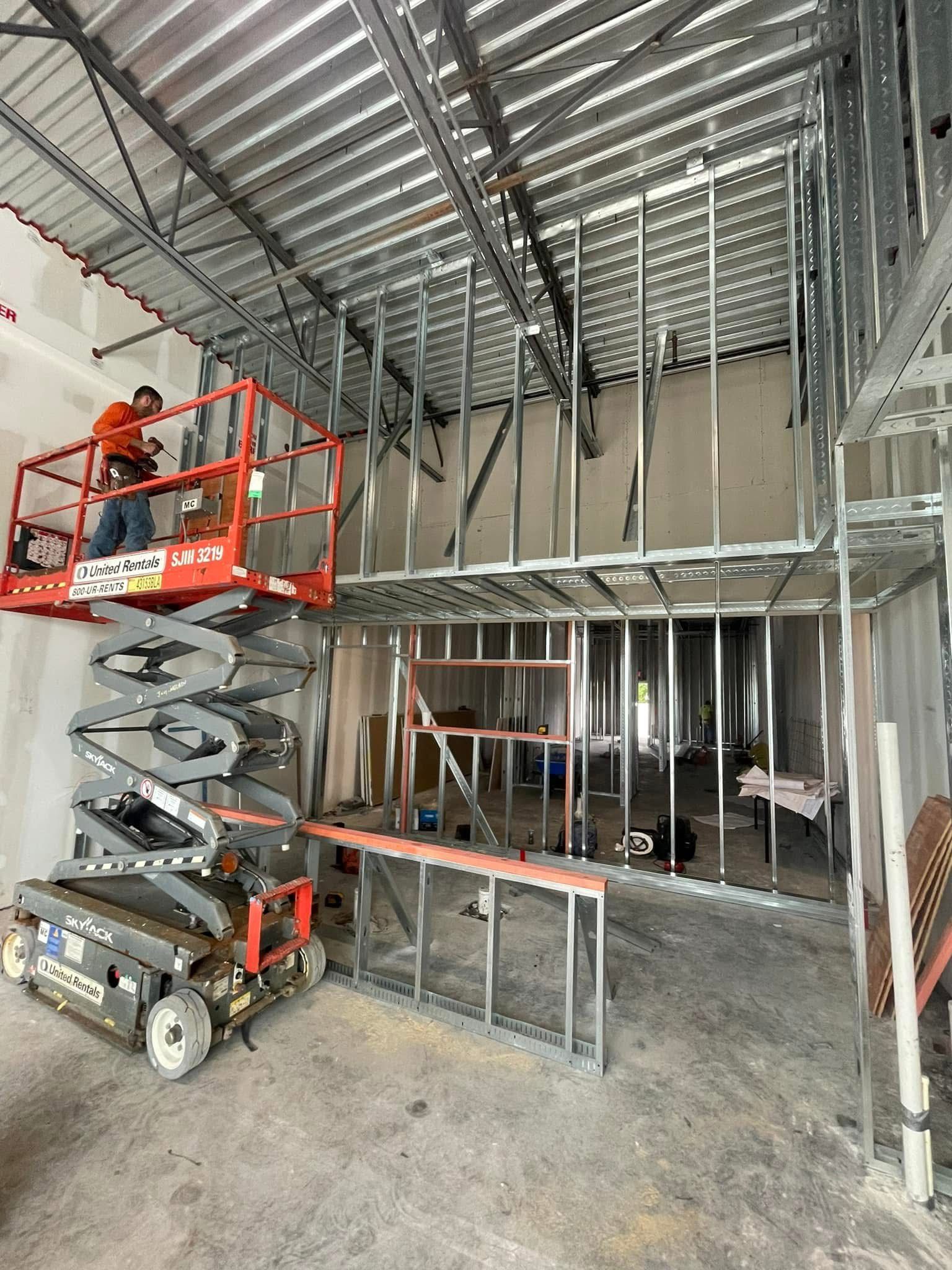 Construction worker on a scissor lift, framing metal studs for a two-story interior. Steel beams and concrete floor.