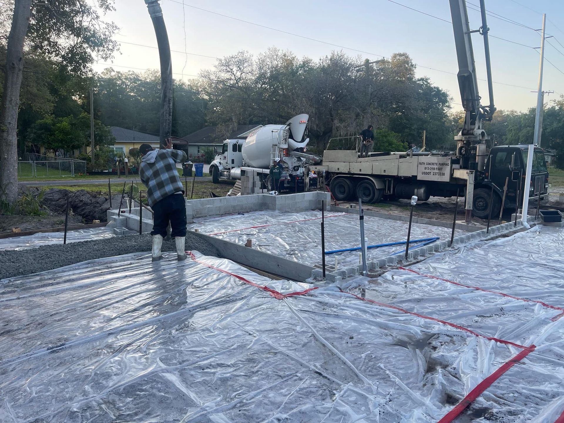 Construction site: worker leveling concrete from pump truck, with foundation and truck visible.