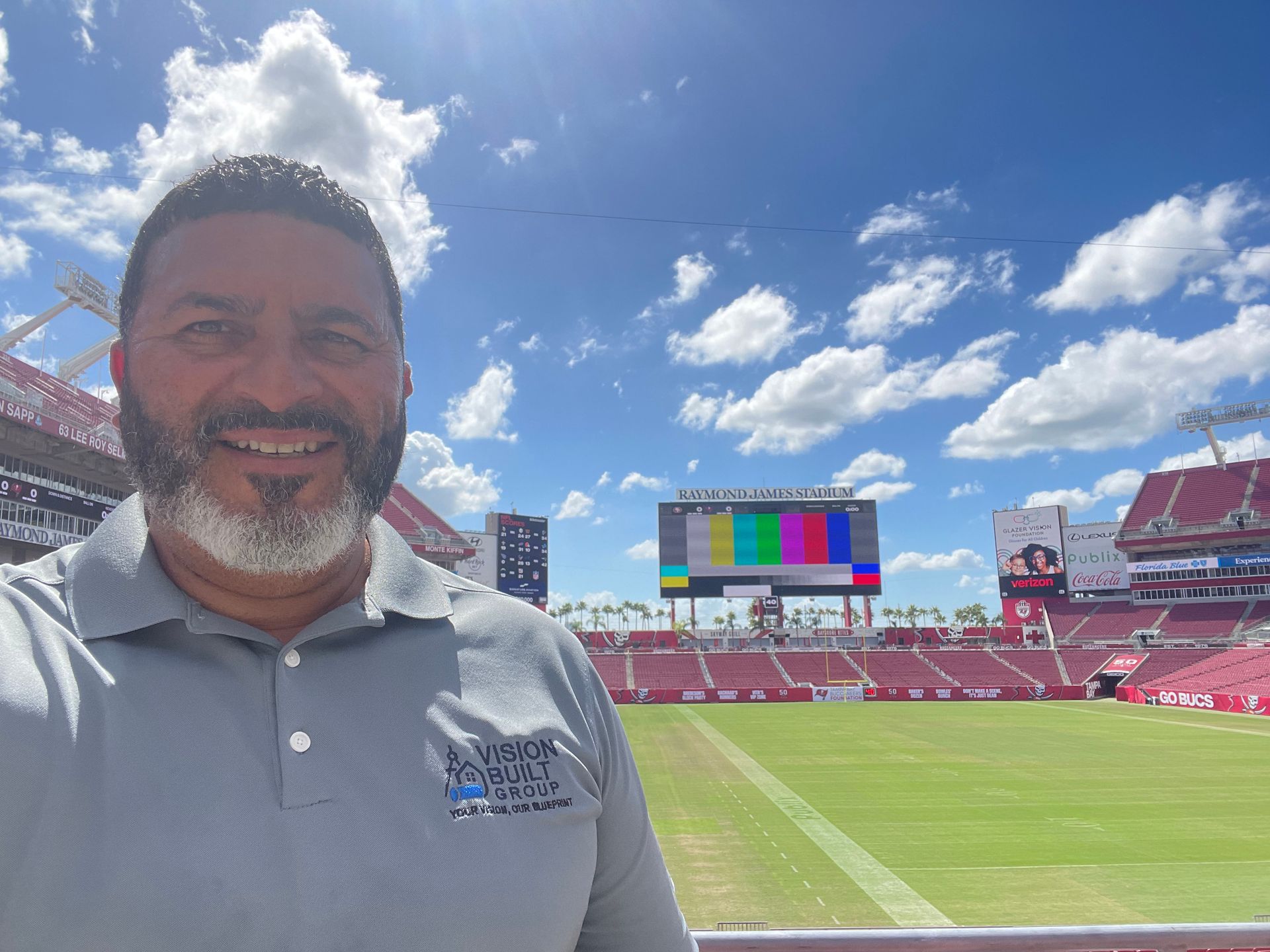 Man in grey shirt smiles in front of a stadium with a green field and a colorful screen under a blue sky.