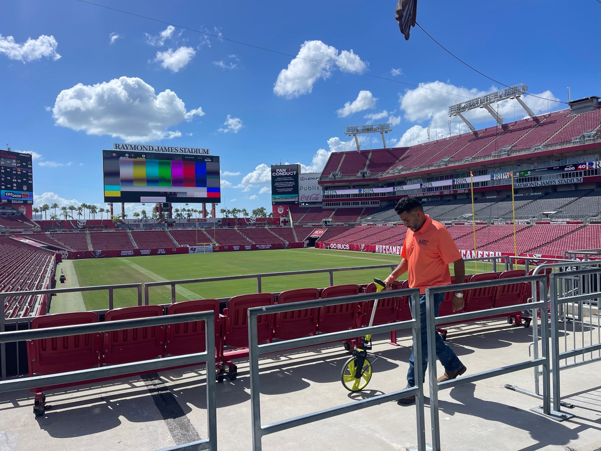 Man measures distance in stadium with a wheel. Red seats, green field, and a large video screen in background.