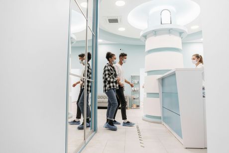 Two people wearing masks and shoe covers walk into a clinic reception area with a lighthouse-themed design.