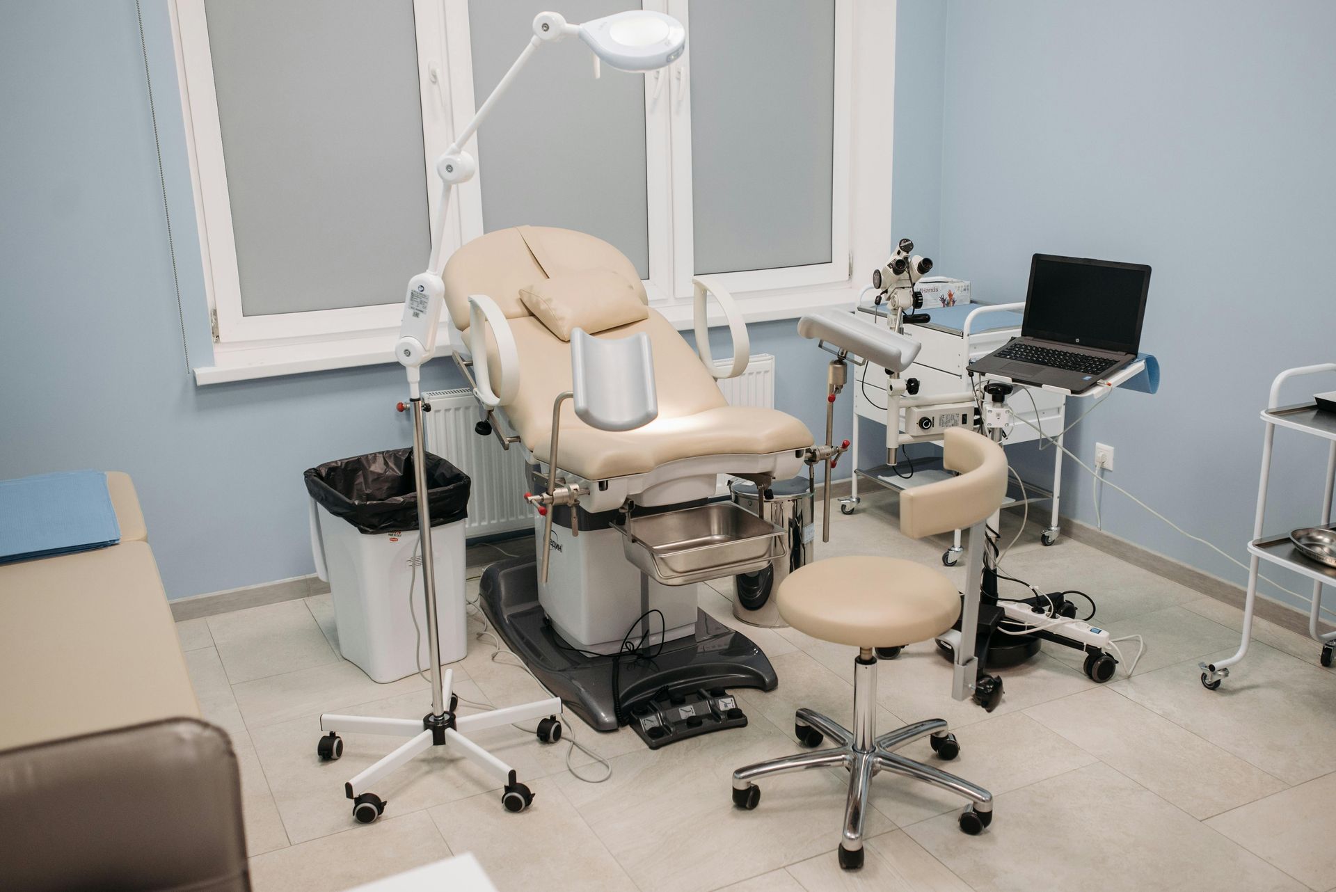 Medical examination room with exam table, light, stool, and equipment. Blue walls and large window.